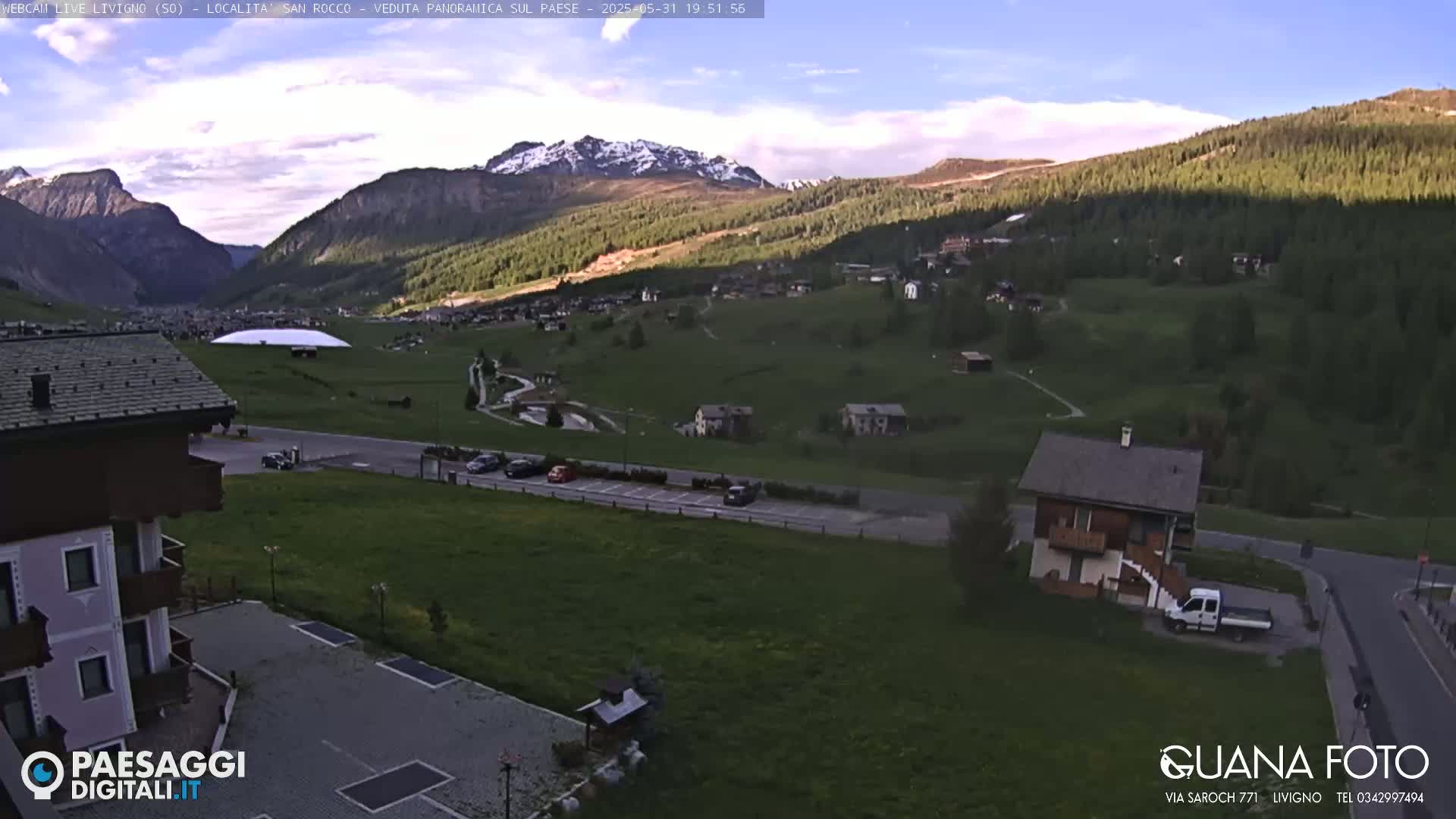 A partly sunny view of a valley town nestled among green hills and mountains, some of which are snow-capped.
