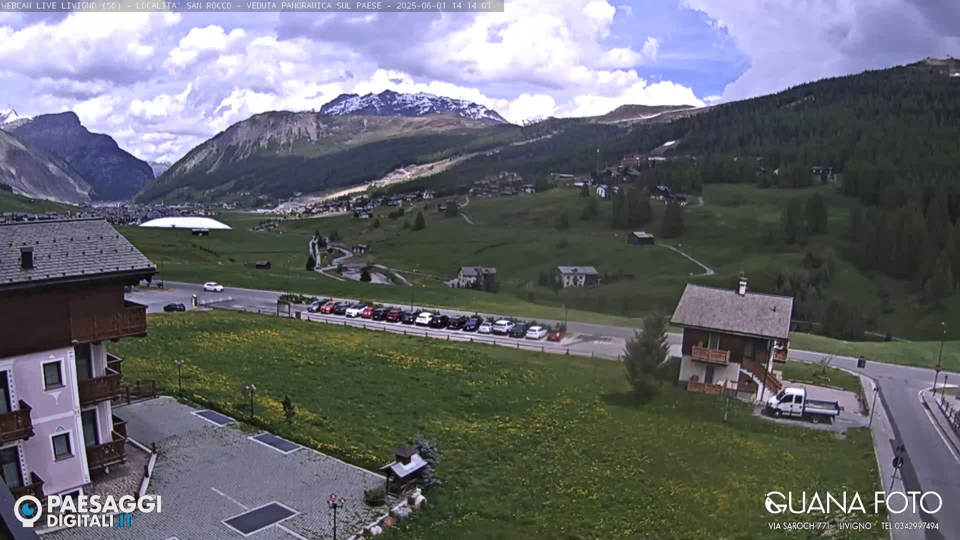 A partly cloudy day reveals a mountain valley town with a row of cars parked near a small house and a larger building.
