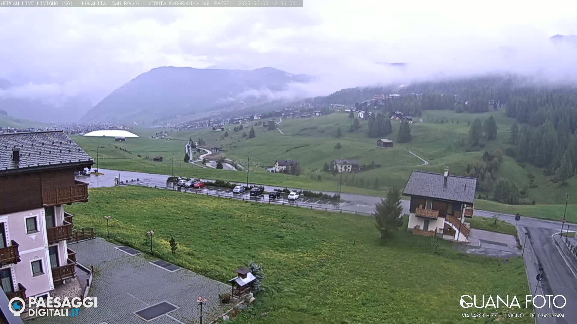 A misty, overcast view of a mountain village with green fields, a parking lot, and several buildings, including a small house and a larger building with balconies.