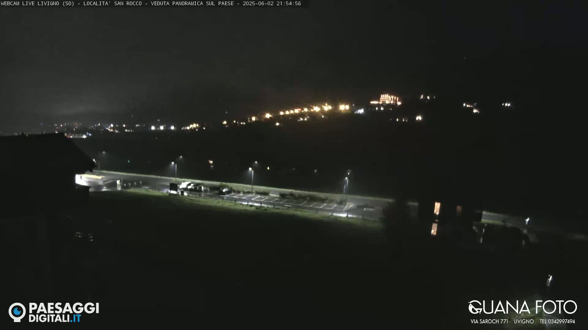 A nighttime long shot of a town nestled on a hill, illuminated by lights, with a dark, empty parking lot and a building in the foreground.
