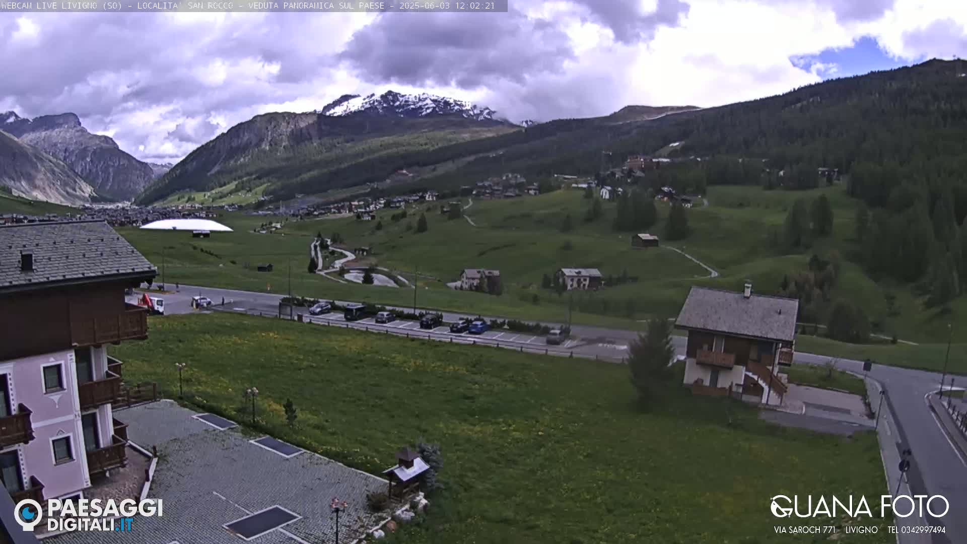 A partly cloudy day reveals a mountain valley town with green fields, a parking lot, and several buildings, including one with a peaked roof.
