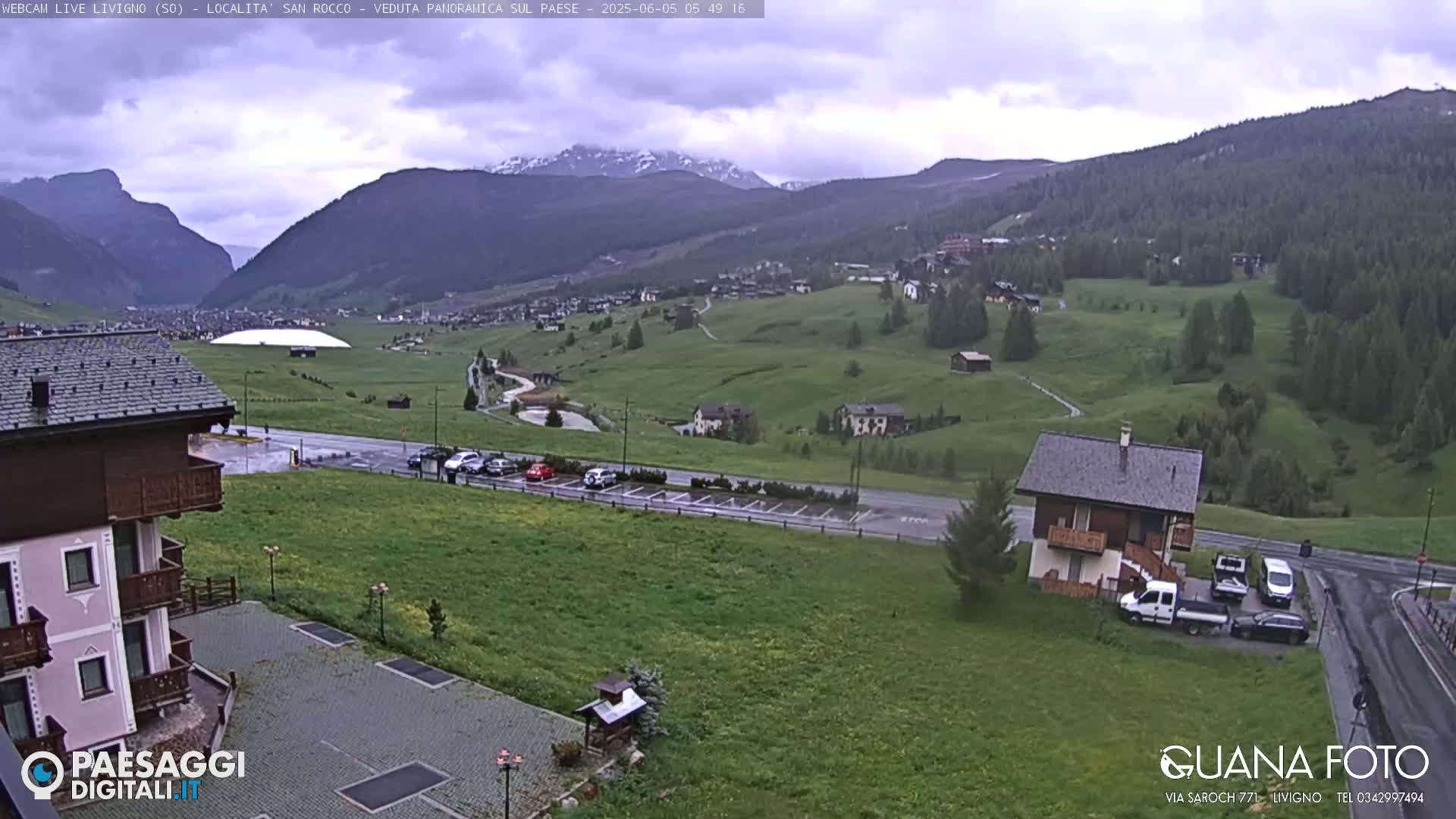 A mountain village with green fields and a few buildings is visible under an overcast sky, with snow-capped mountains in the background.