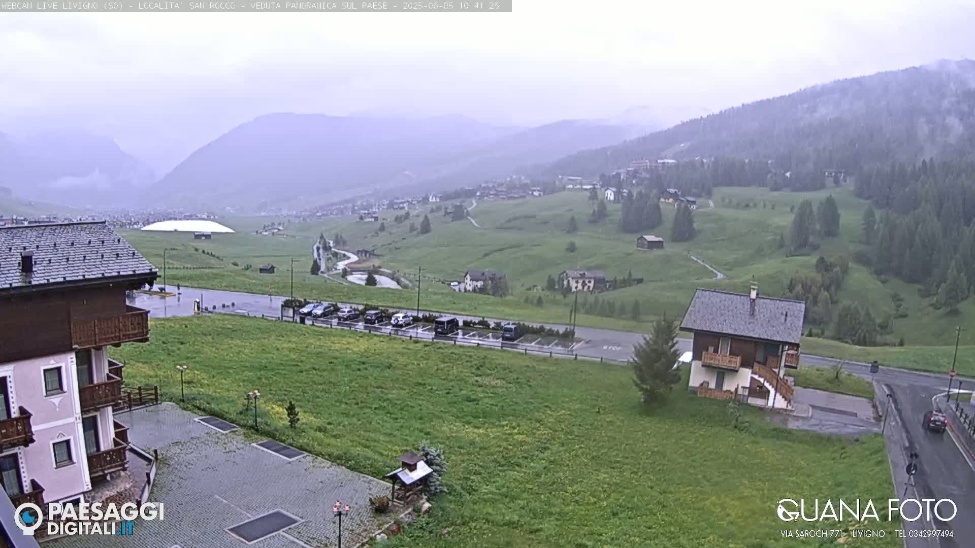 A rainy view of a mountain village with green hills, a parking lot, and buildings, including a large white dome-like structure in the distance.