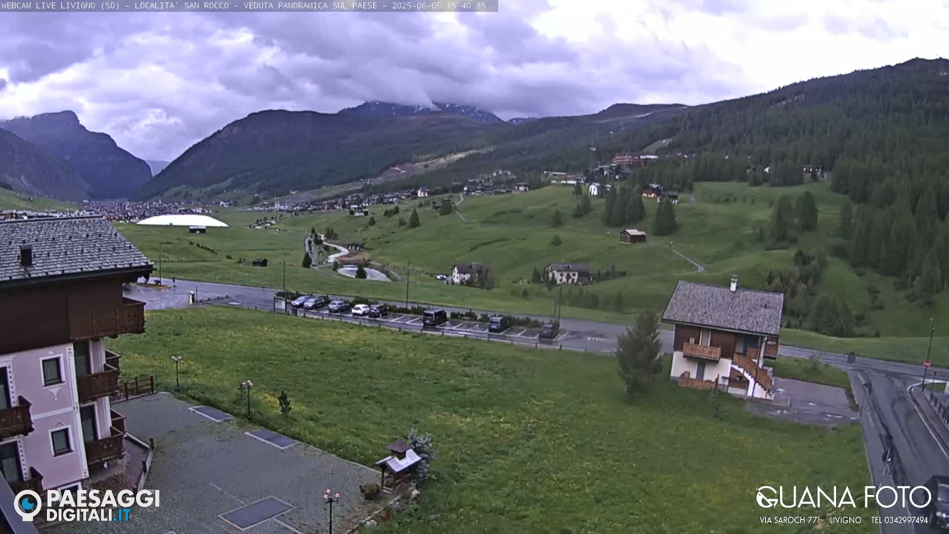 A view of a mountainous village under a cloudy sky, showing green hills, buildings, a parking lot, and a small house.