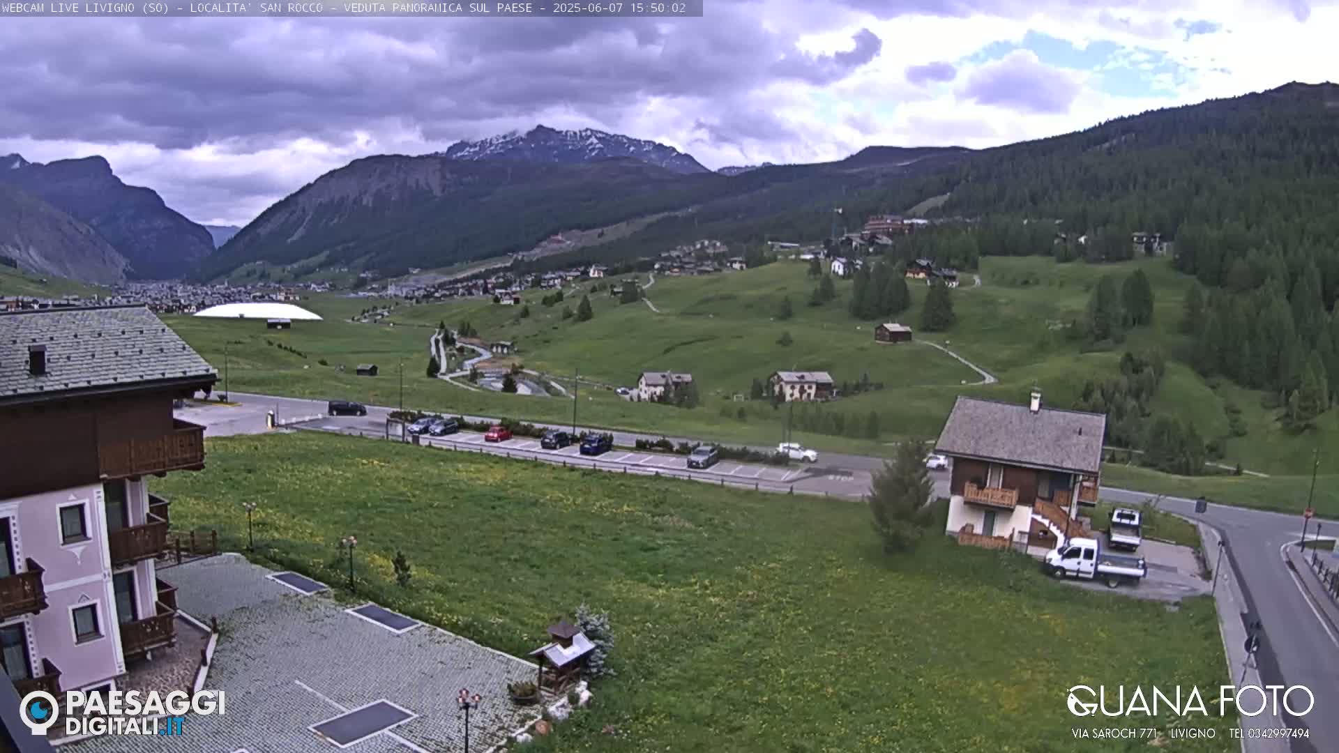 A mountain village nestled in a valley under a partly cloudy sky, with green fields, buildings, and parked cars visible.