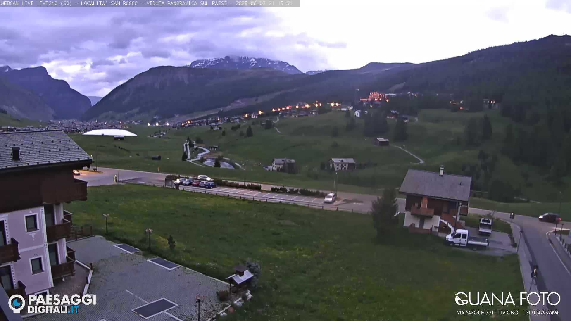A mountain village at dusk under a cloudy sky, with a small building and parking lot in the foreground.