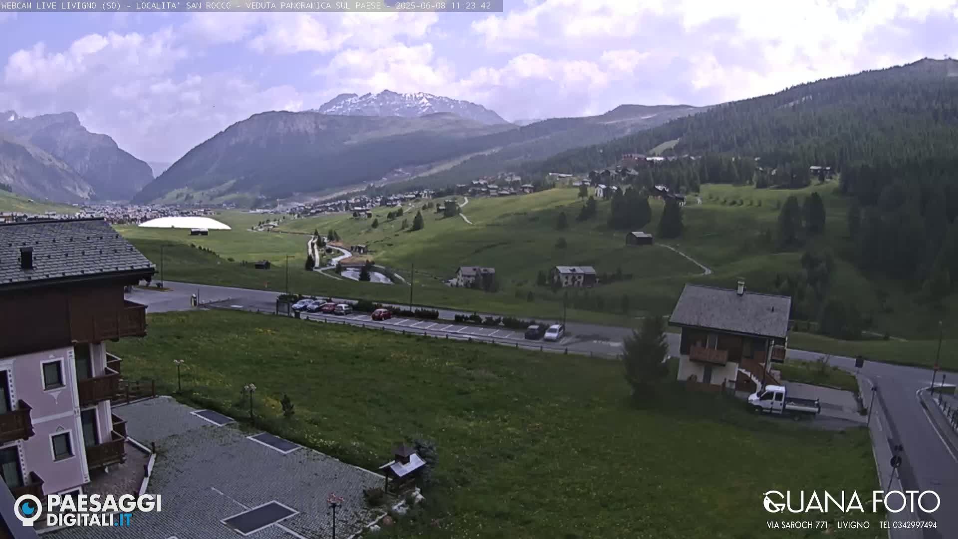A partly cloudy day reveals a mountain valley town nestled among green hills and mountains, with a parking lot and buildings visible in the foreground.