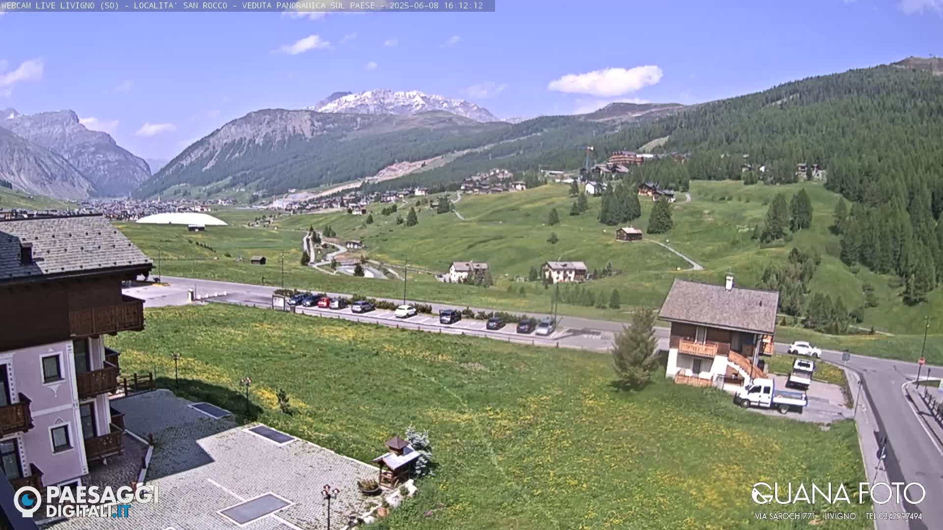 A sunny view of a mountainous village nestled in a valley, with green fields, a parking lot, and several buildings, including a small house.