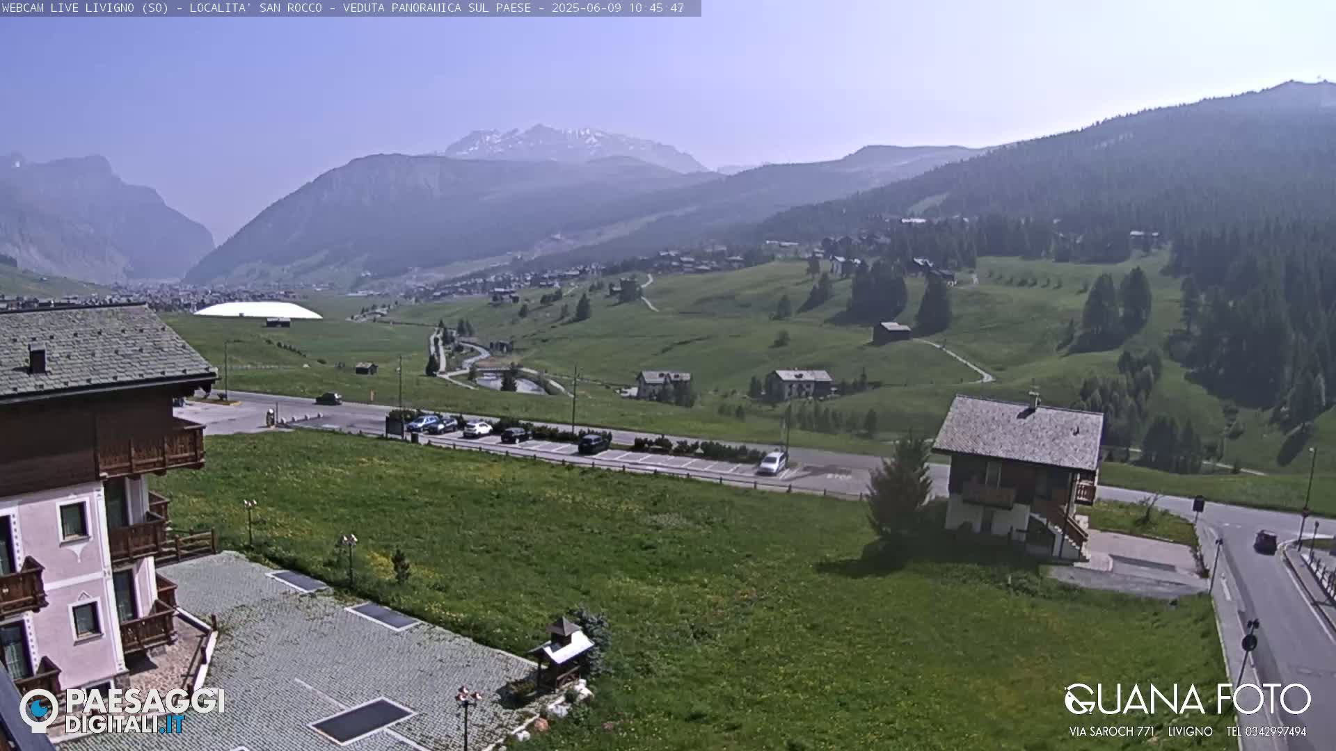 A sunny mountain valley scene shows a village nestled amongst green hills and a parking lot near a building, with distant mountains under a clear sky.