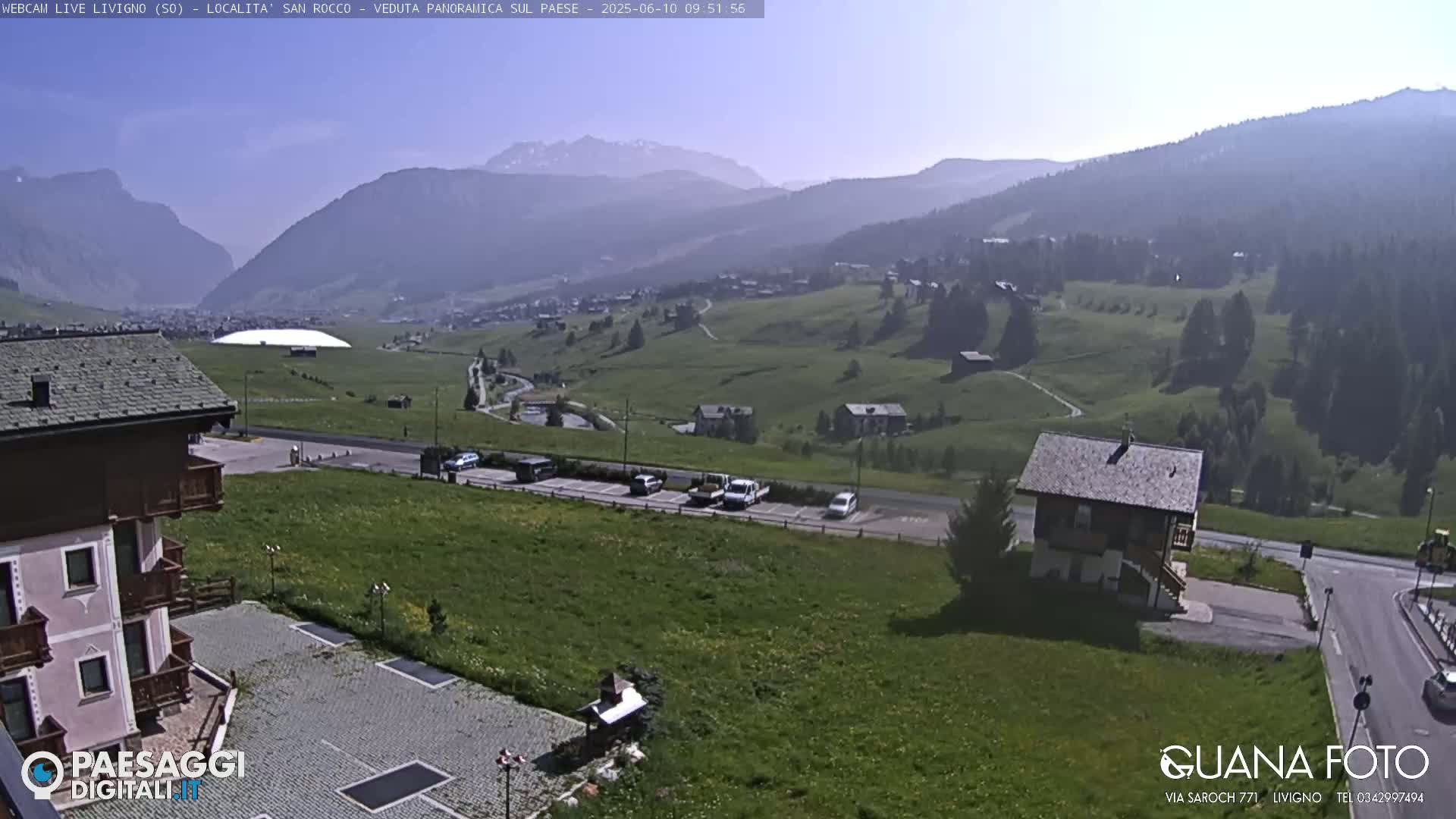 A sunny, hazy mountain vista overlooks a small village with a parking lot and a few buildings, including a chalet-style house.