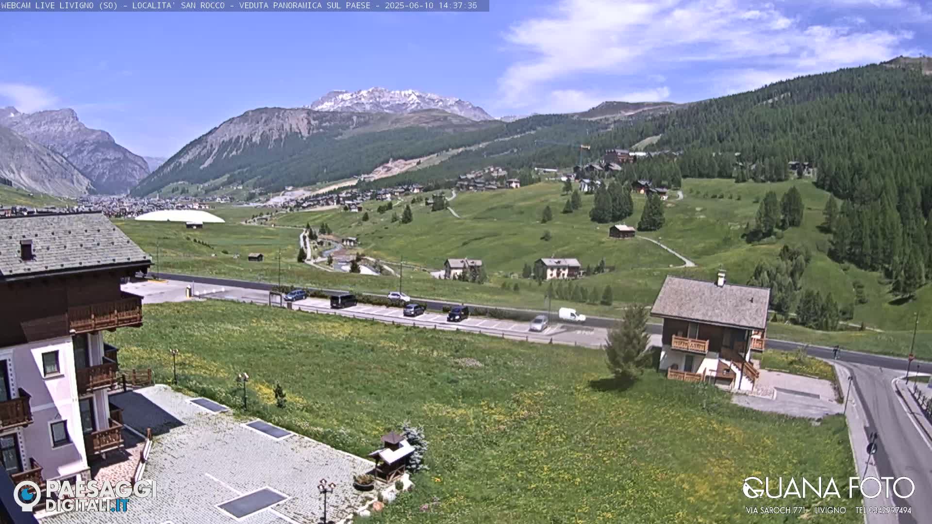 A sunny mountain village scene shows green hills, a parking lot with several cars, and a small brown house, with snow-capped mountains in the background.