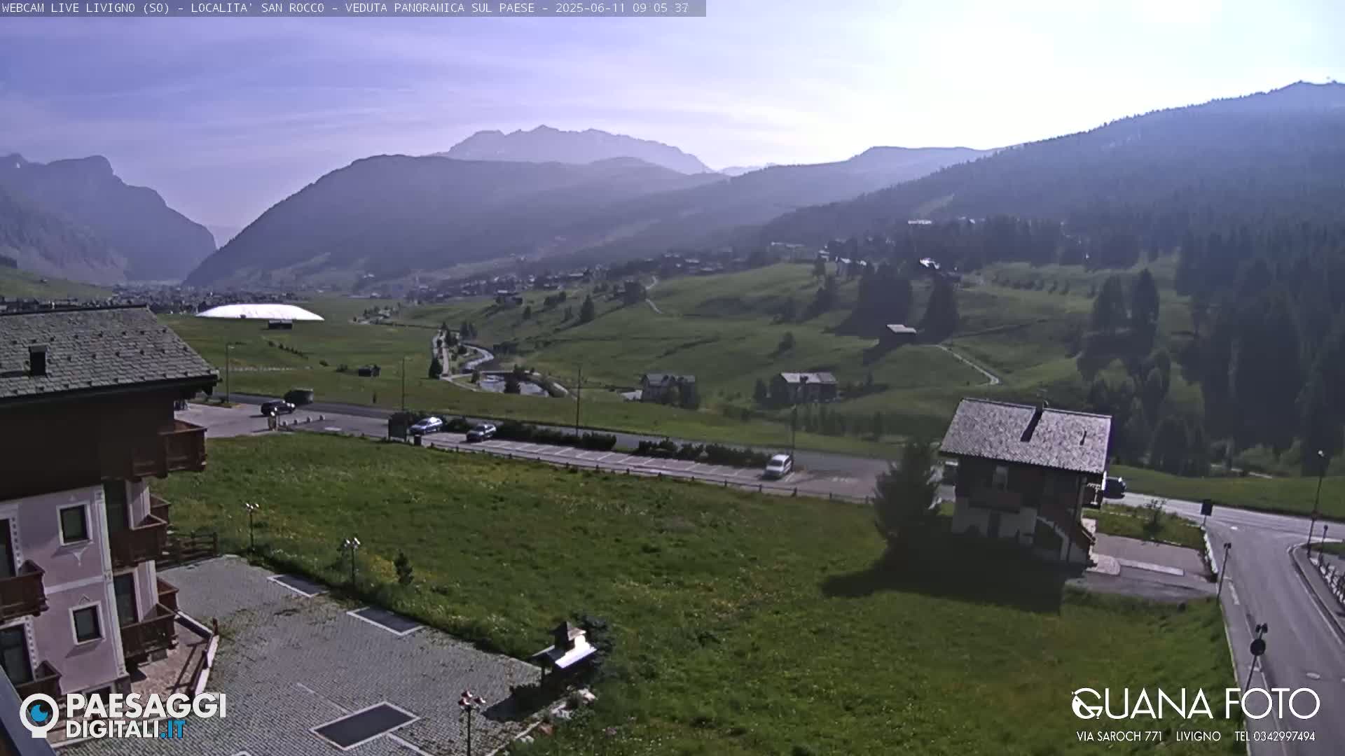 A mountain valley scene on a sunny day, showing green hills, a few buildings, and a road with parked cars.