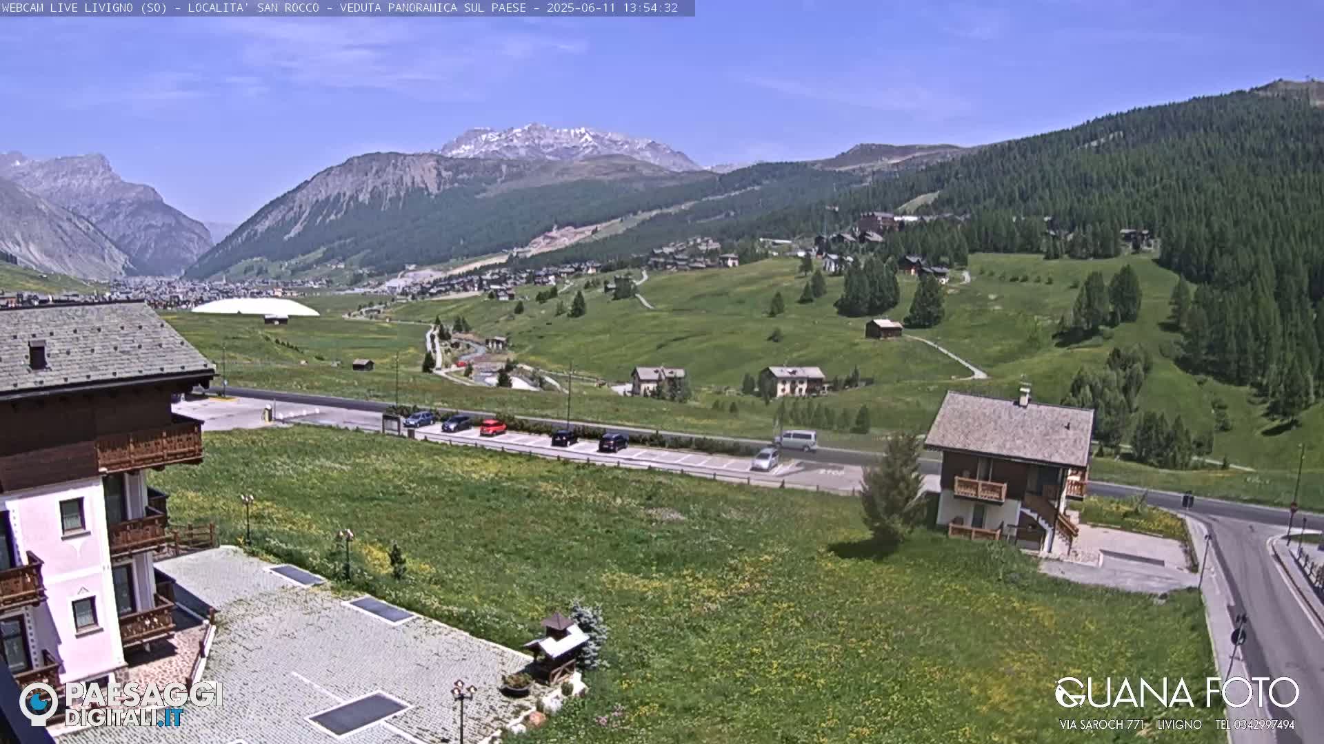 A sunny mountain valley scene shows a small village nestled among green hills, with snow-capped mountains in the background and a few cars parked near buildings.