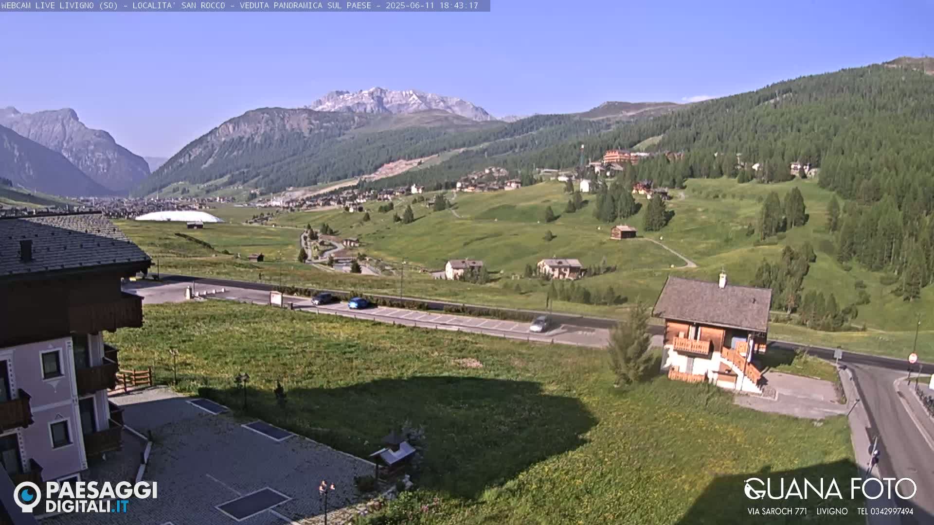 A sunny, panoramic view of a mountainous valley with a village nestled amongst green fields and a few cars parked along a road.