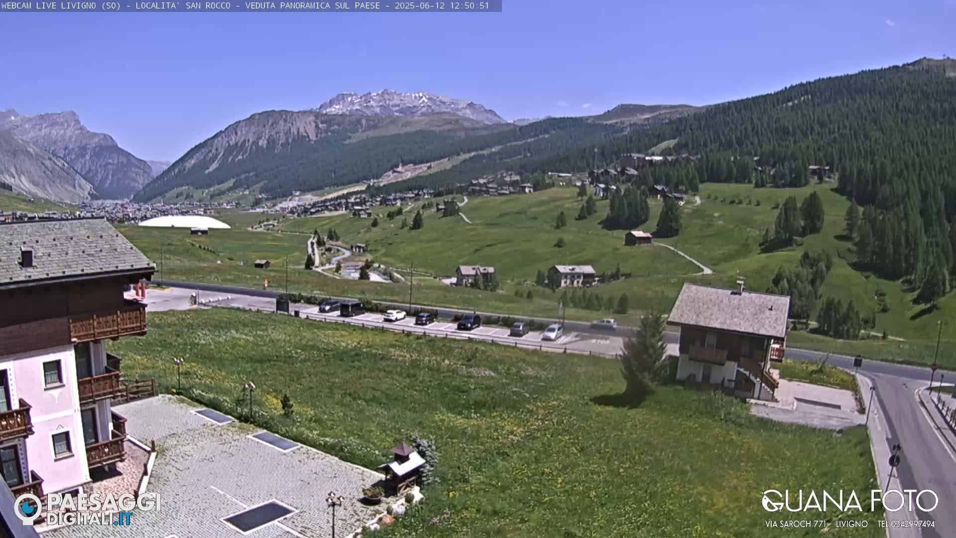 A sunny view of a mountainous region shows a village nestled in a green valley, with several buildings, cars in a parking lot, and a large white dome-like structure in the distance.