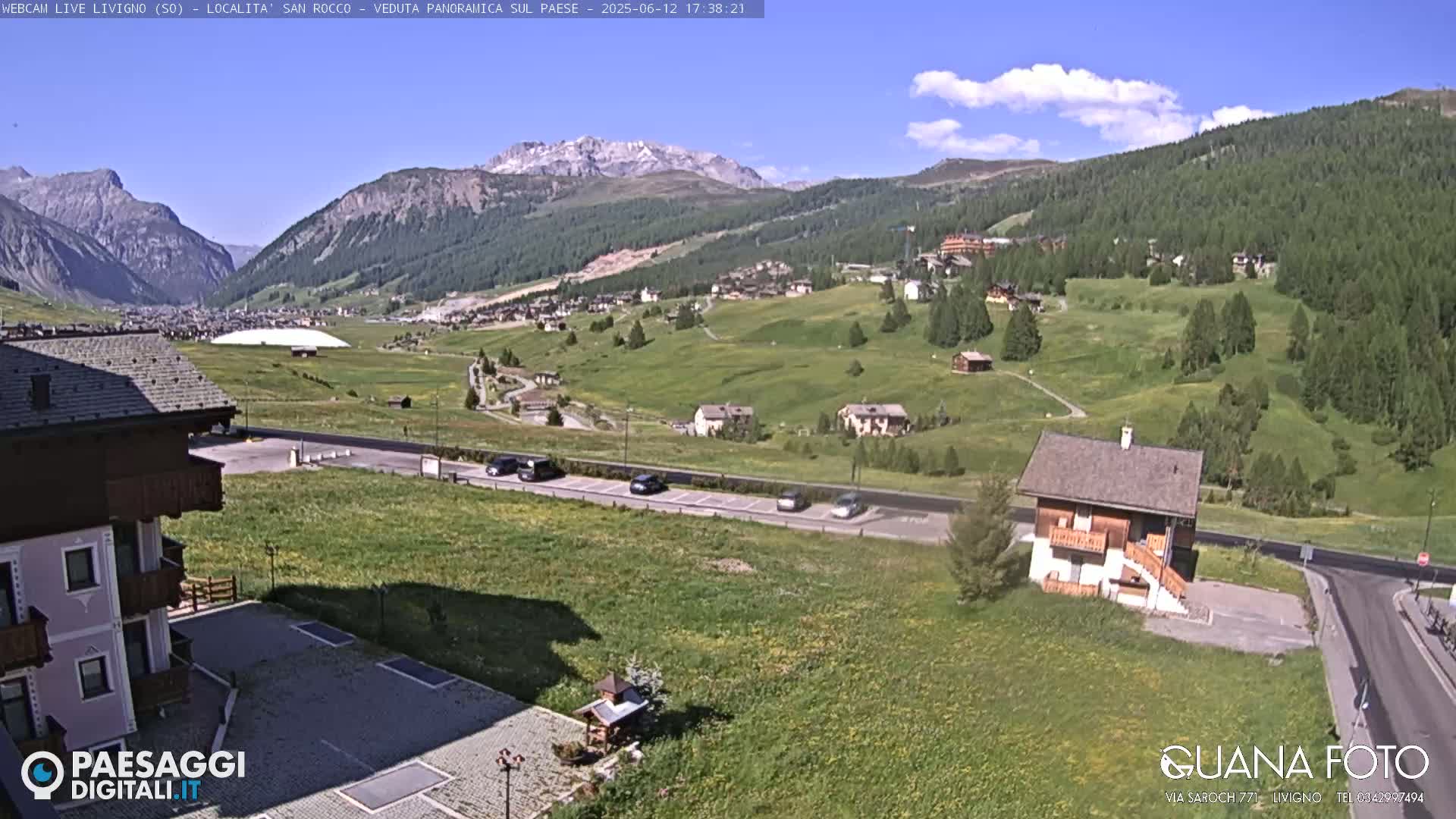 A sunny day reveals a mountain valley with green fields, a few buildings, and parked cars, under a partly cloudy sky.