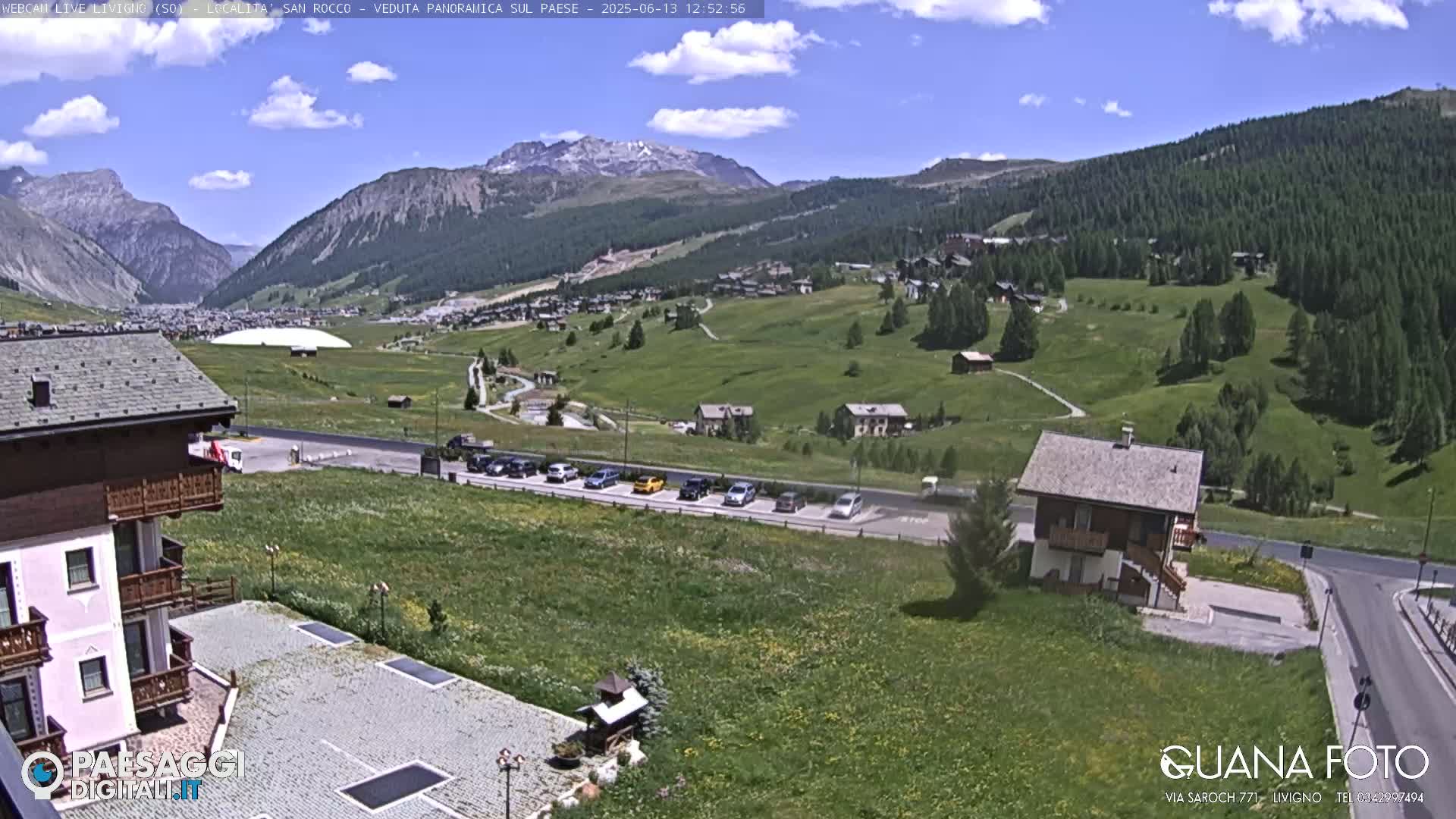 A sunny, mountainous landscape with green fields, buildings, and parked cars; a large structure resembling a dome is visible in the distance.