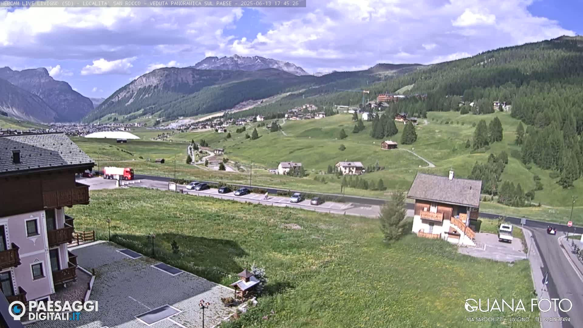 A partly cloudy day reveals a mountain village nestled in a green valley, with a parking lot and several buildings visible in the foreground.