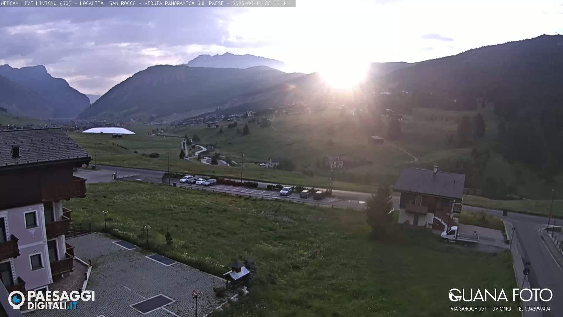 A partly cloudy day reveals a mountain village nestled in a green valley, with a parking lot and several buildings visible in the foreground.