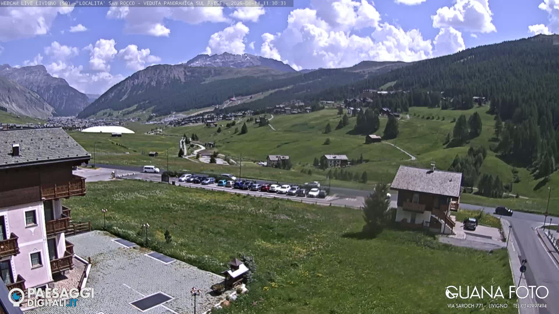 A sunny day in a mountainous region shows a village nestled in a valley, with cars parked in a lot and several buildings visible, including a large structure resembling a dome.