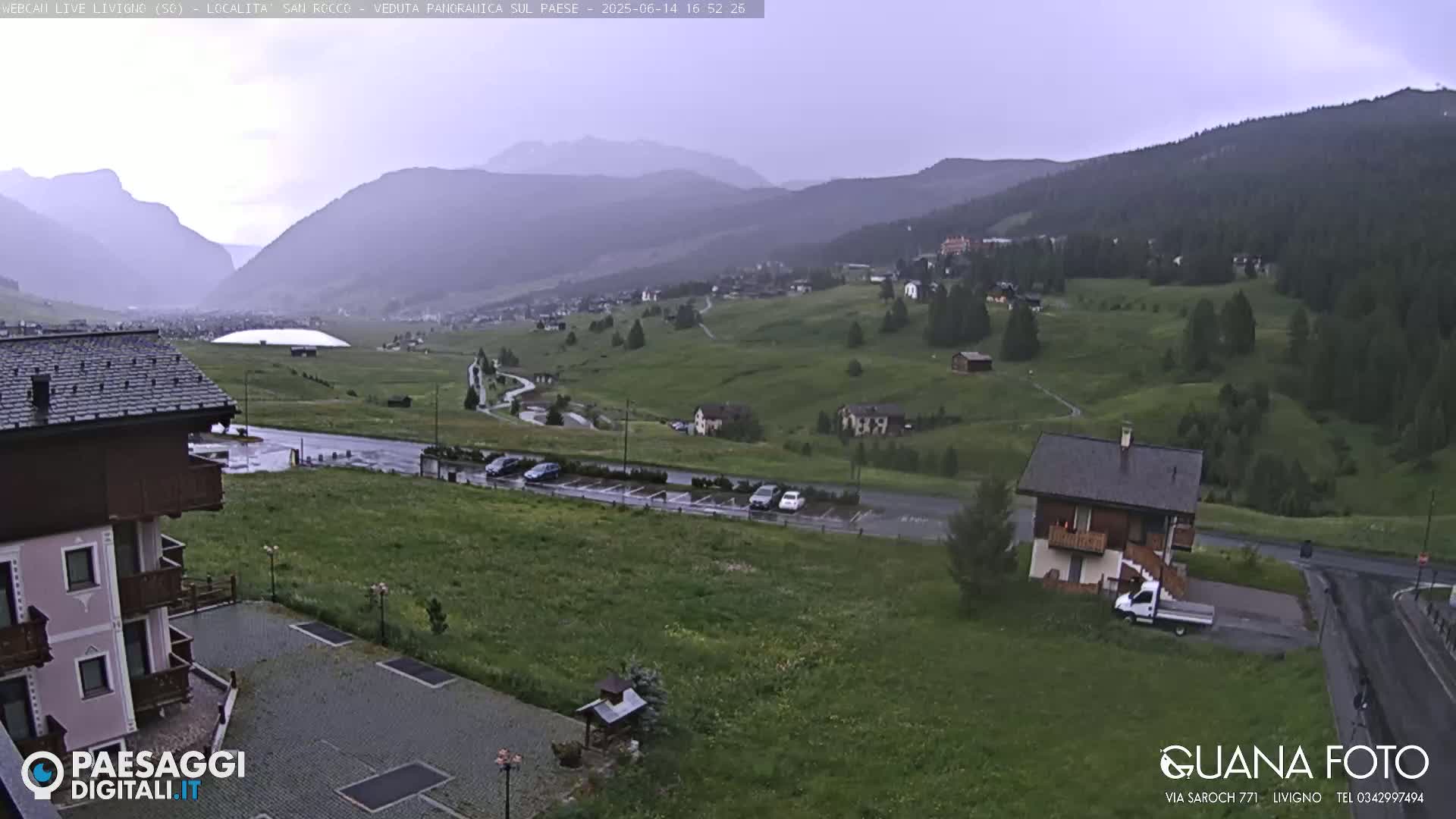 A rain-soaked mountain valley village scene with a few cars parked in a lot, a small house, and a larger building visible in the foreground, all set against a backdrop of rolling green hills and distant mountains under an overcast sky.