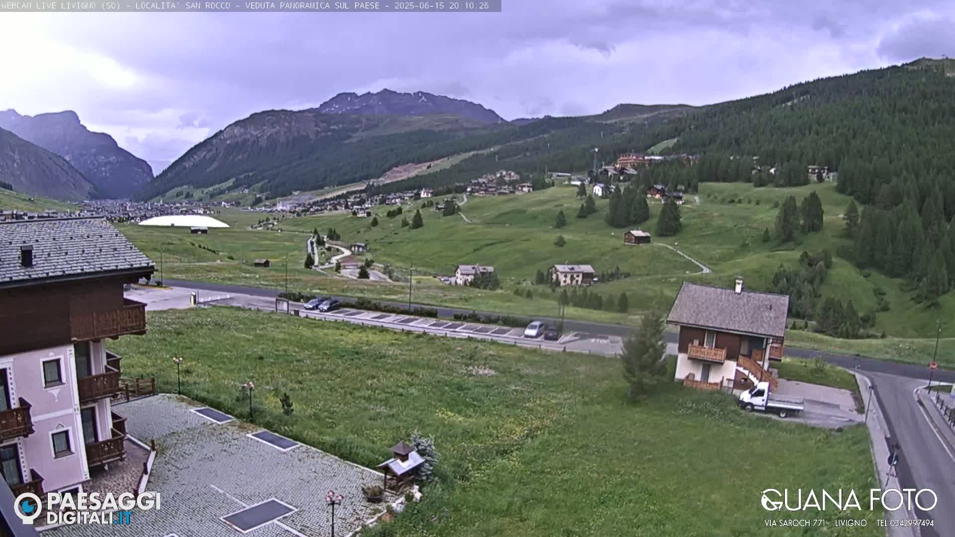 A partly cloudy day reveals a mountain valley town with green hillsides, a parking lot, and several buildings, including a small house near the foreground.