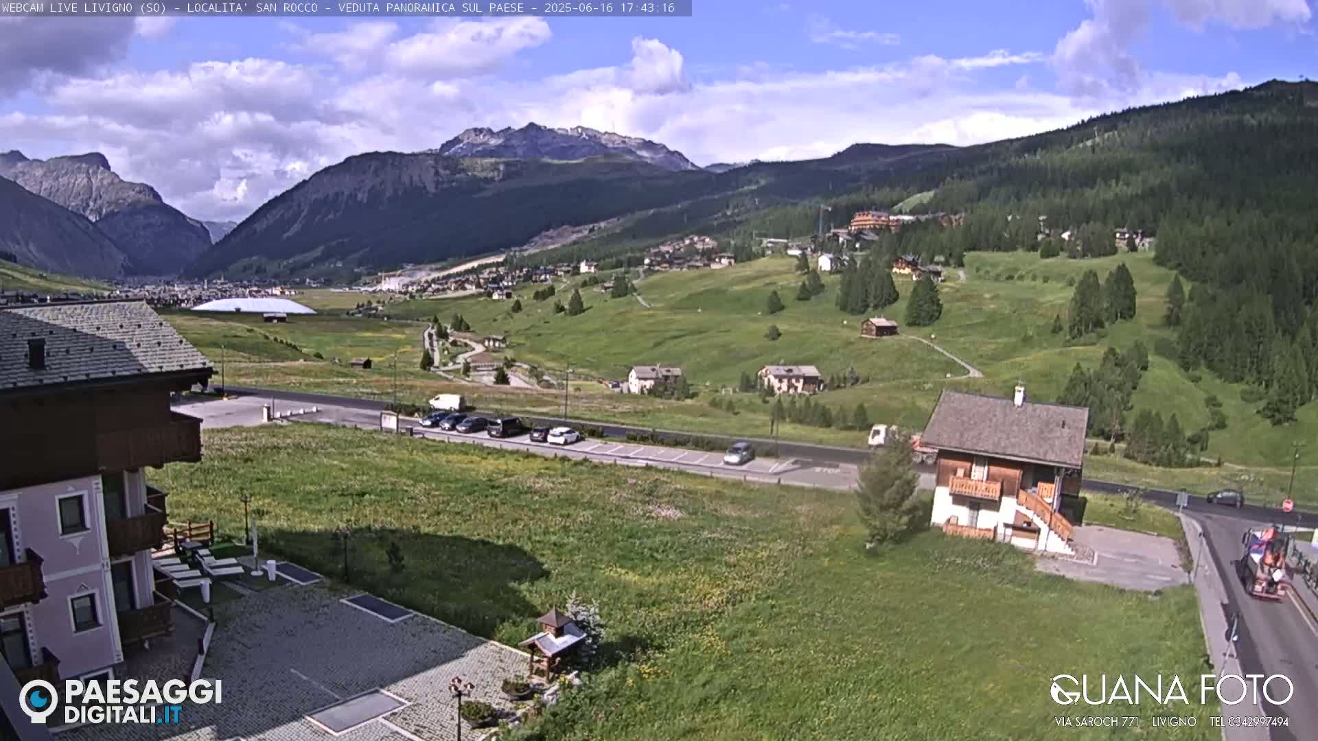 A sunny mountain valley scene with green fields, a few buildings, parked cars, and a road, under partly cloudy skies.