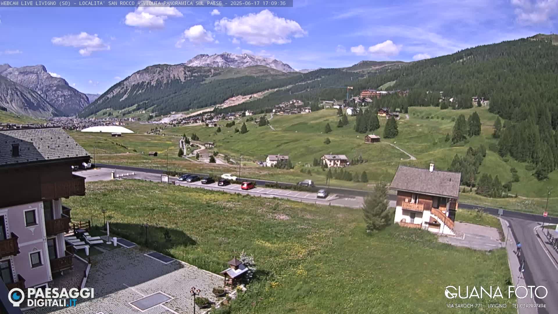 A sunny day in a mountainous region shows a village nestled in a valley, with cars parked near a small house and a larger building in the foreground, under a partly cloudy sky.