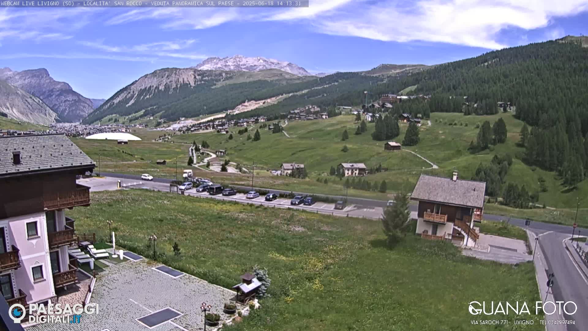 A sunny mountain valley scene shows a small village nestled among green hills, with a snow-capped mountain peak in the distance and several cars parked near a small house.