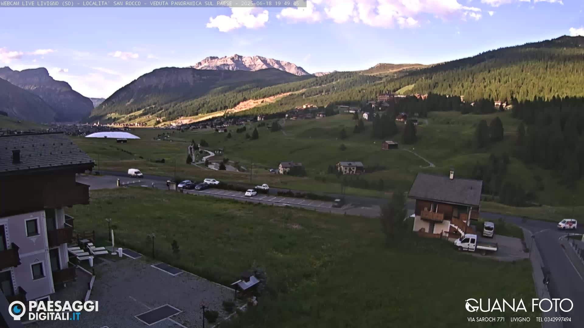 A mountain village nestled in a valley under a partly cloudy evening sky, with a few cars parked near a small house and a larger building.