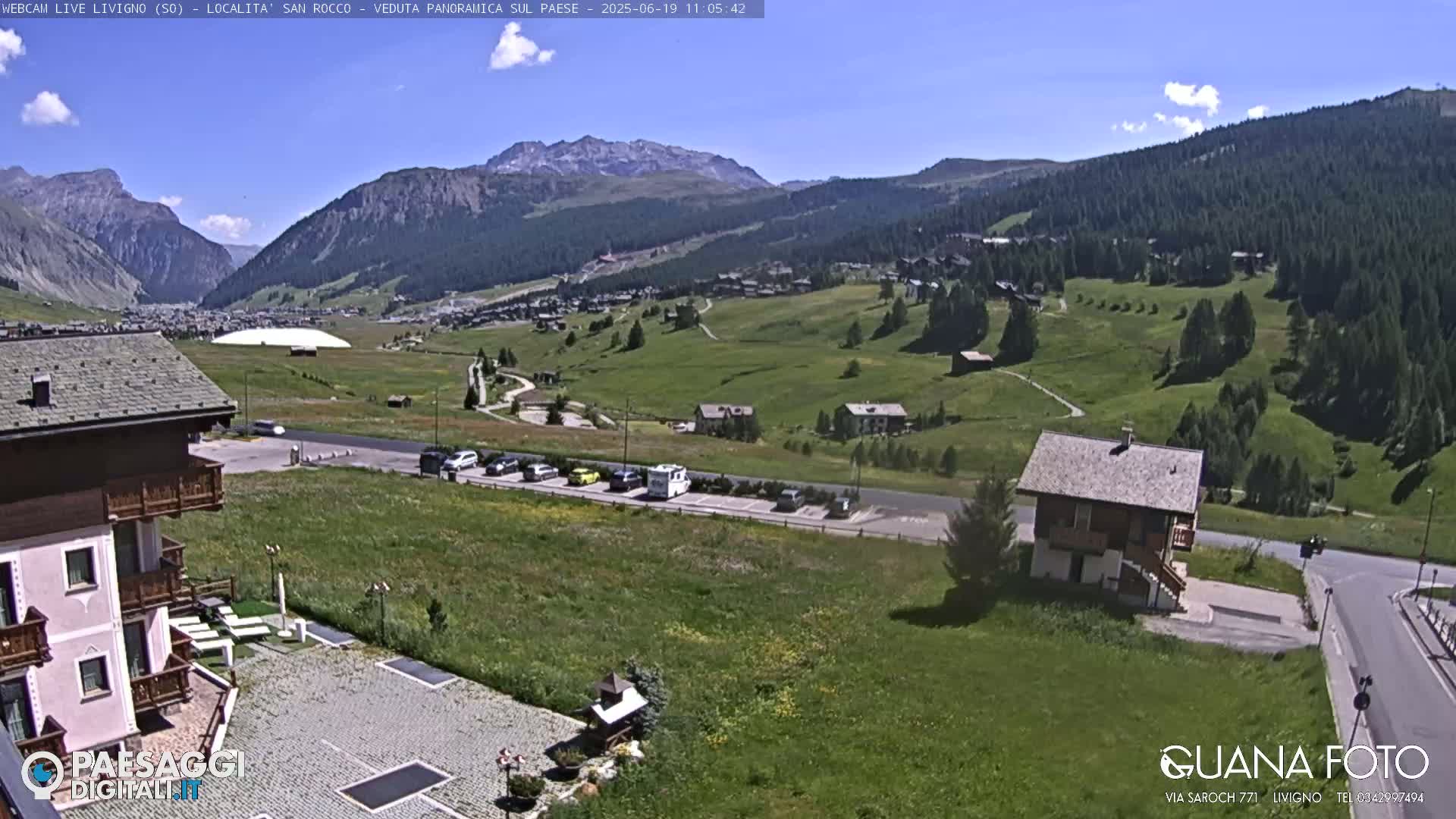 A sunny mountain village scene shows a grassy valley with buildings, parking lots, and a large white structure in the distance, all set against a backdrop of mountains under a mostly clear blue sky.