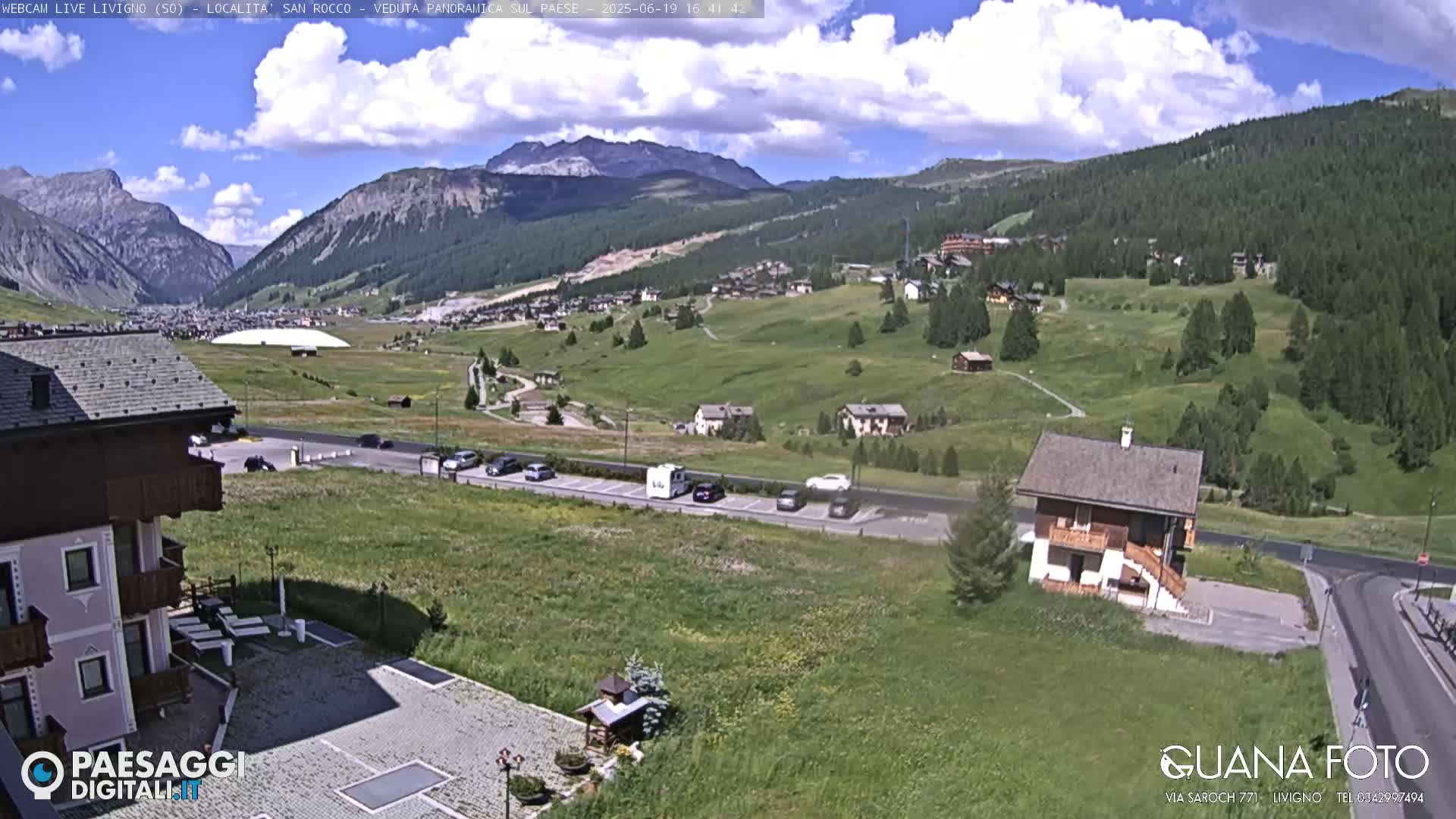 A partly cloudy day reveals a mountain valley town with green hills, a parking lot with vehicles, and several buildings, including a small house near the road.