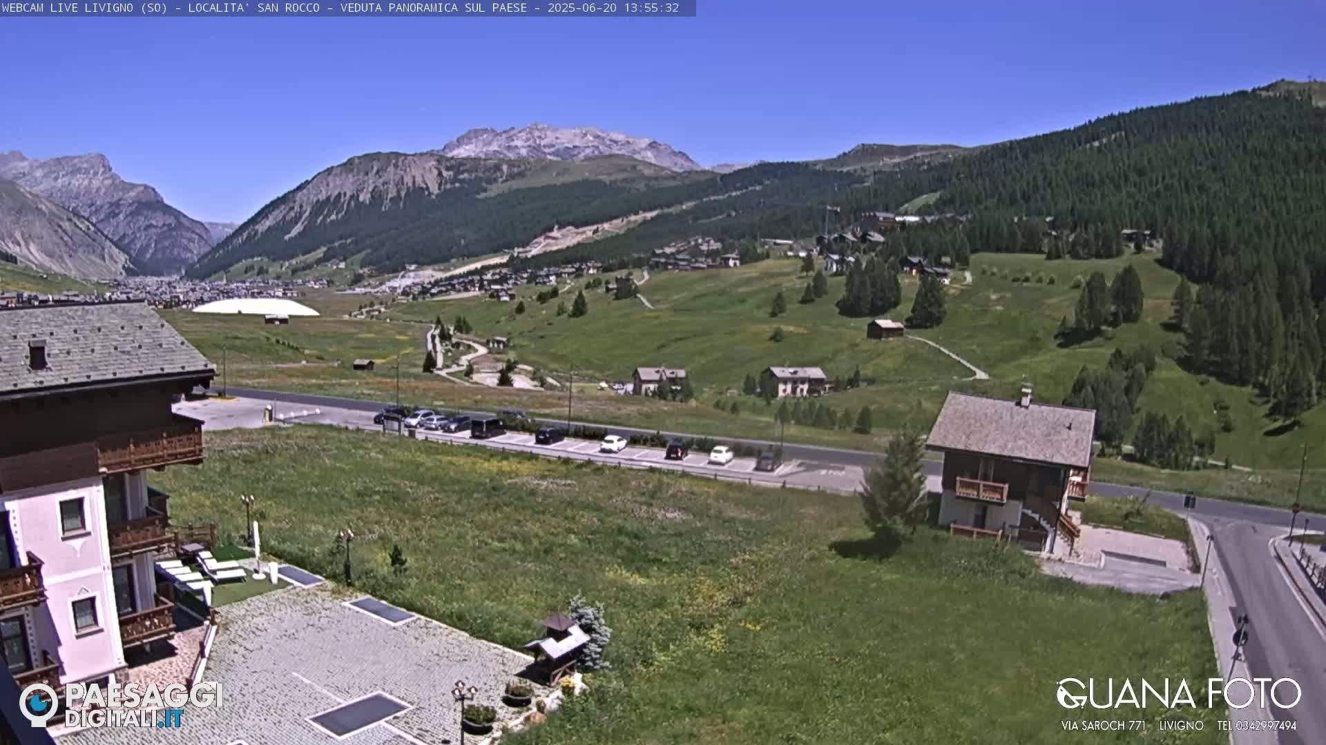 A sunny mountain vista shows a village nestled in a valley with green hillsides and a few cars parked in a lot near a small house, all under a clear blue sky.