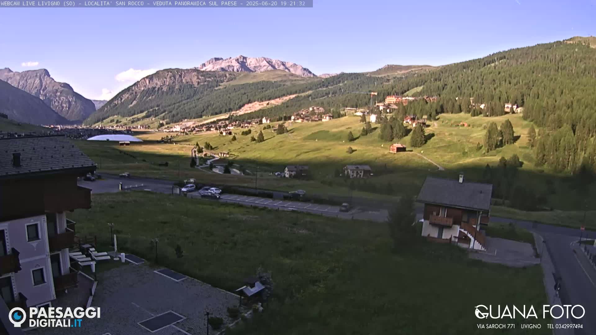 A sunny, high-altitude village nestled in a valley between mountains, with a few cars visible on a road below.