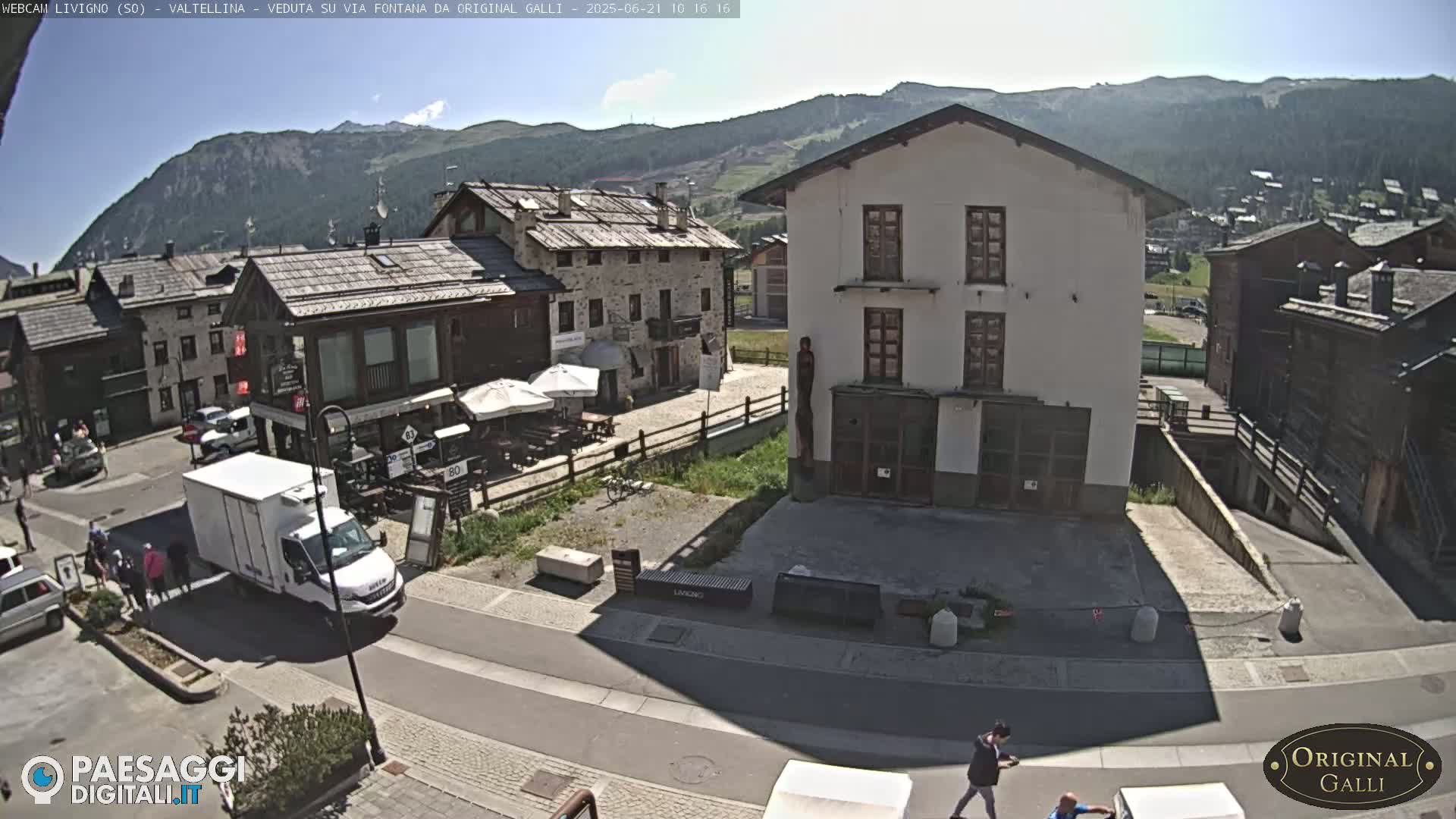 A sunny view of a village nestled in a mountain valley, with several buildings, a street, and a few people walking.