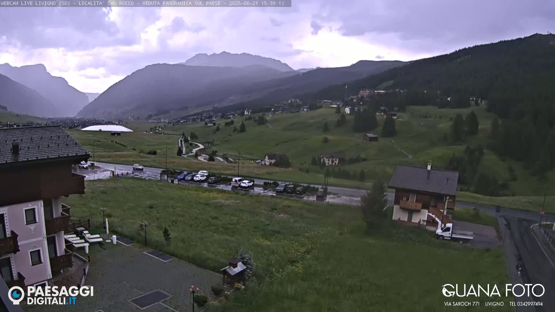 A mountain village in overcast conditions, viewed from a slightly elevated position, showing a parking lot, buildings, and a large grassy area.