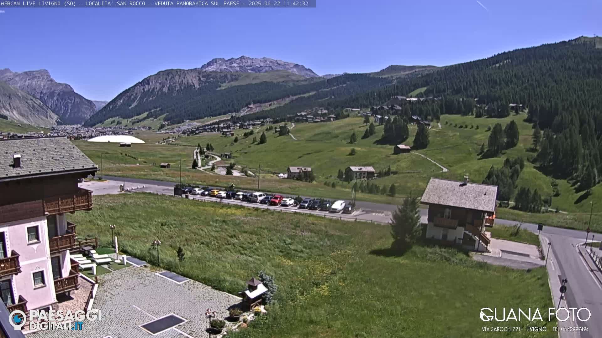 A sunny, panoramic view of a mountainous village with green hills, a parking lot filled with cars, and several buildings, including a chalet-style house.