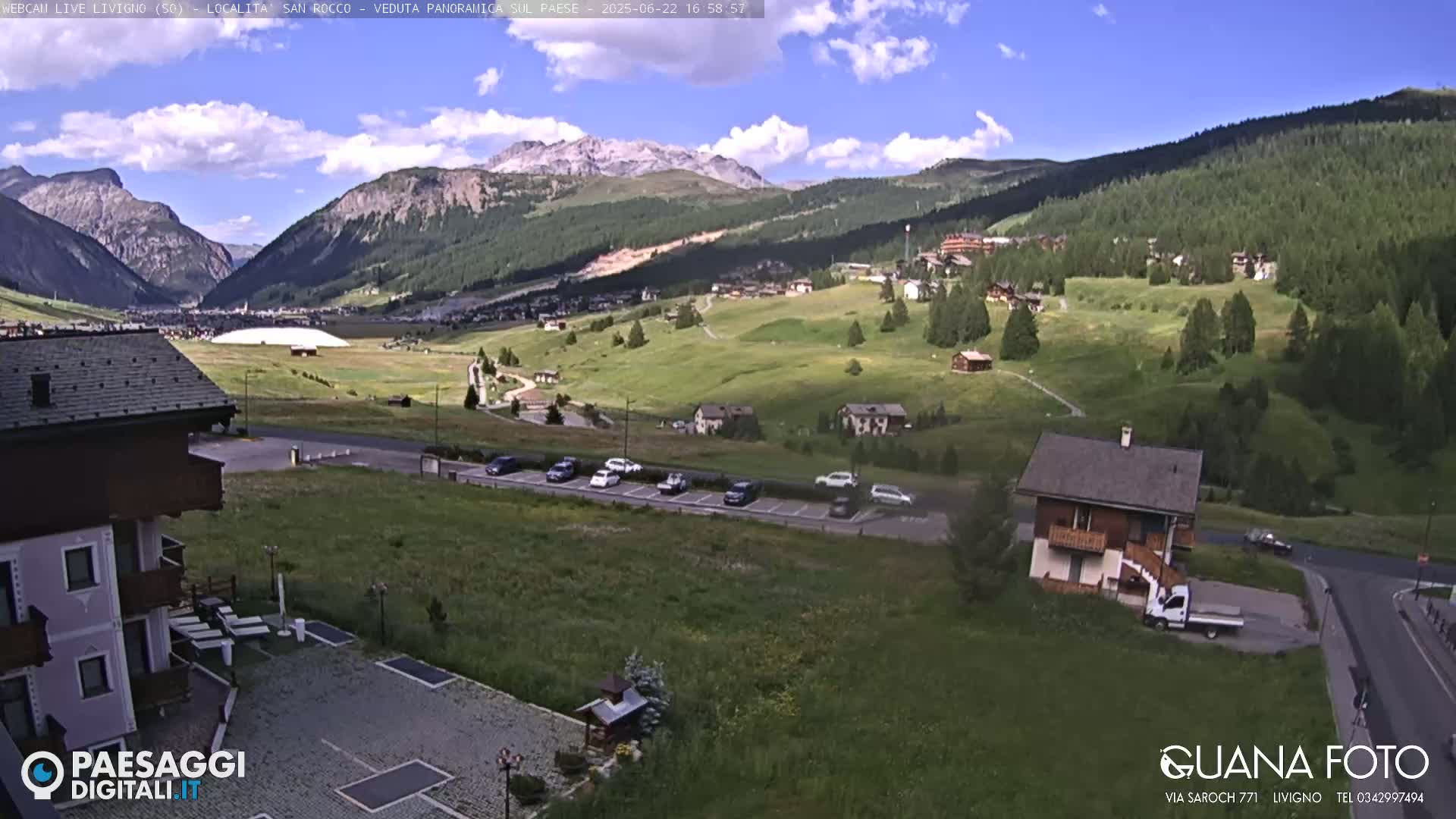 A partly cloudy day reveals a mountain valley town with several cars parked in a lot, a few buildings, and green rolling hills under a mostly clear blue sky.