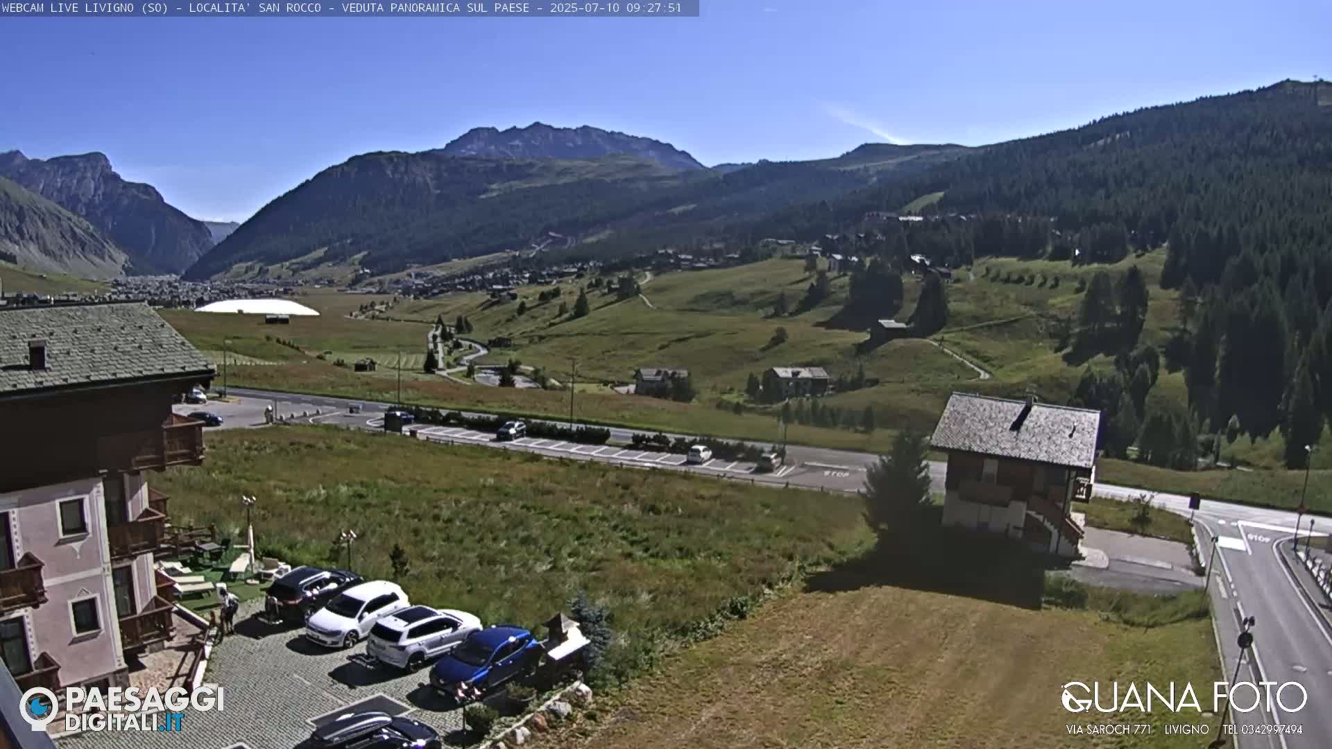 A sunny day reveals a mountain valley town with cars parked near buildings, and green hills and mountains in the background.