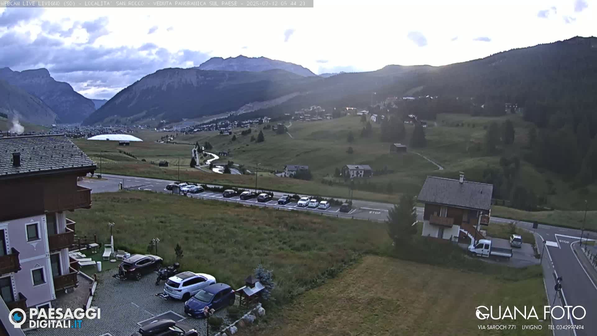 A sunny day reveals a mountain valley town with cars parked near buildings, and green hills and mountains in the background.