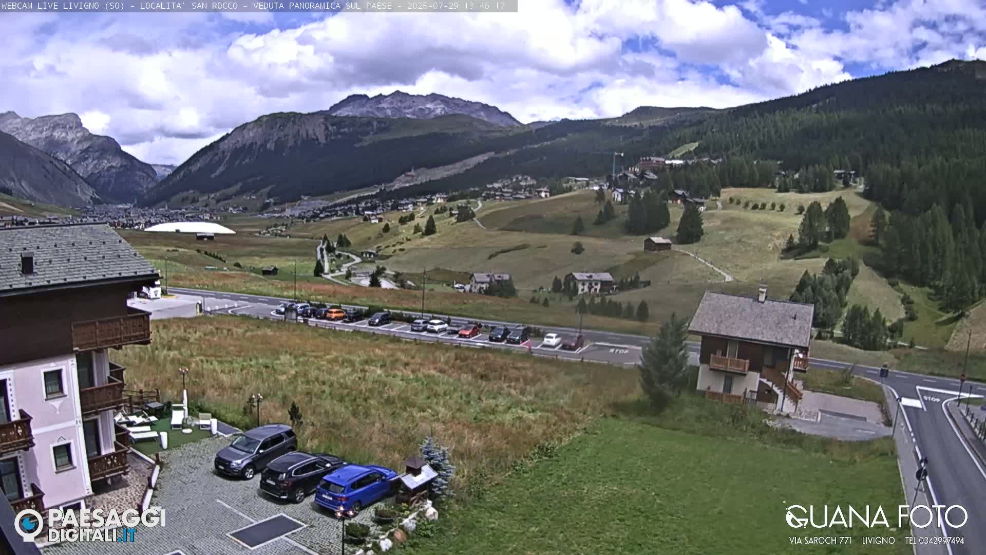 A sunny, high-altitude view of a village nestled in a valley surrounded by mountains, with a parking lot and several buildings visible in the foreground.