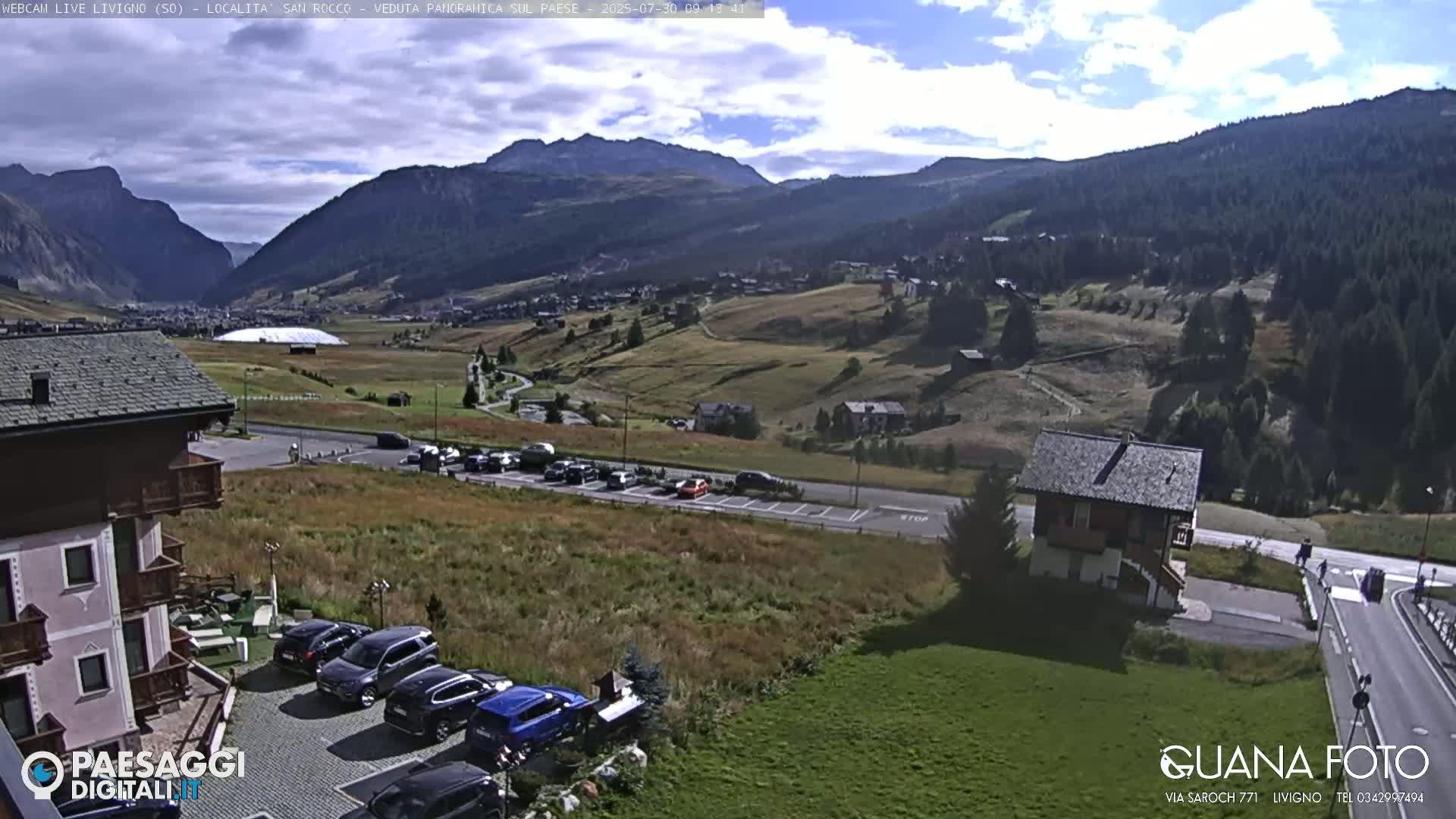A mountain valley with a partly cloudy sky, showing a village, parking lots with cars, and buildings, including a chalet-style house.