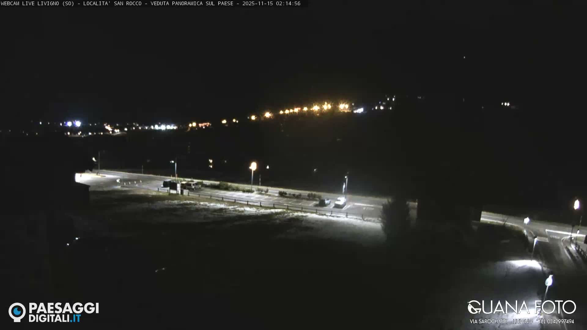 A nighttime panoramic view captures a sparsely lit road and parking lot with a light dusting of snow or frost on the ground, leading to a distant illuminated town under a dark sky.