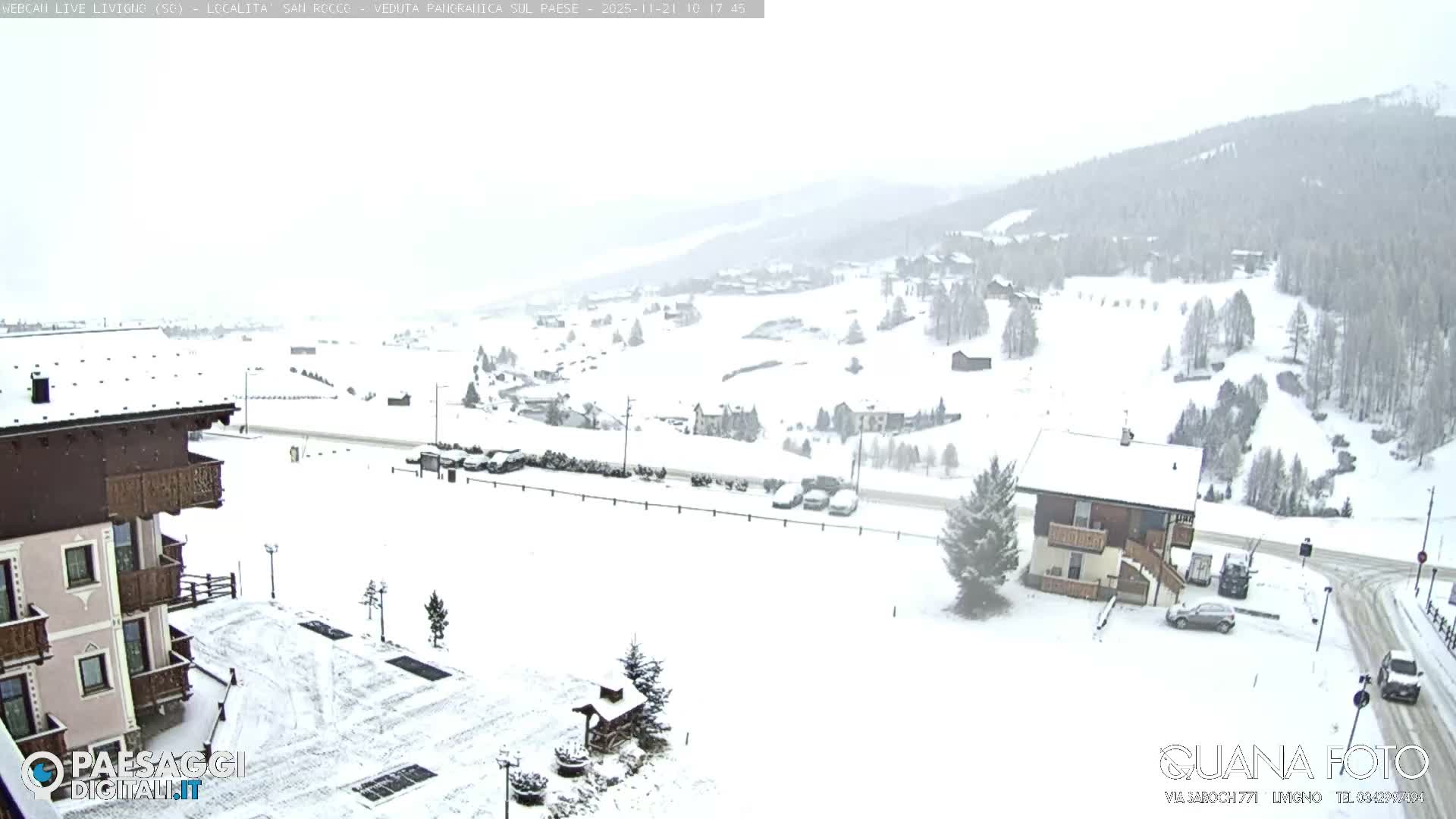 A wide view of a snow-covered mountain village features numerous buildings, roads with a few vehicles, and snow-dusted evergreen trees under a bright, overcast sky.