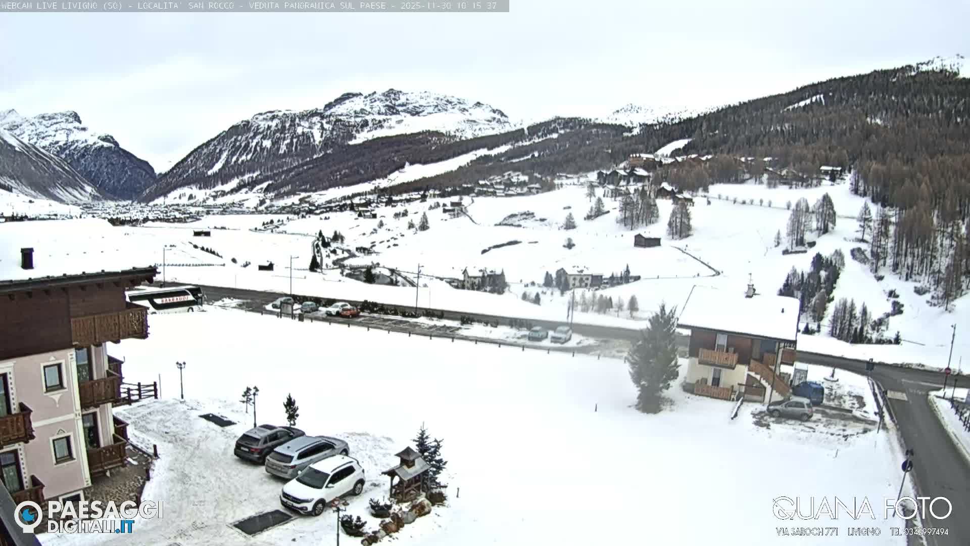 A panoramic winter scene displays a snow-covered mountain village with numerous buildings, roads, and trees, nestled between forested peaks under an overcast sky.