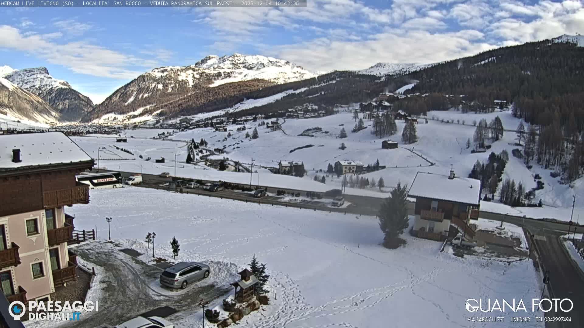 The image presents a panoramic view of a snow-covered mountain valley dotted with buildings and trees, with vehicles on clear roads, under a partly cloudy sky.