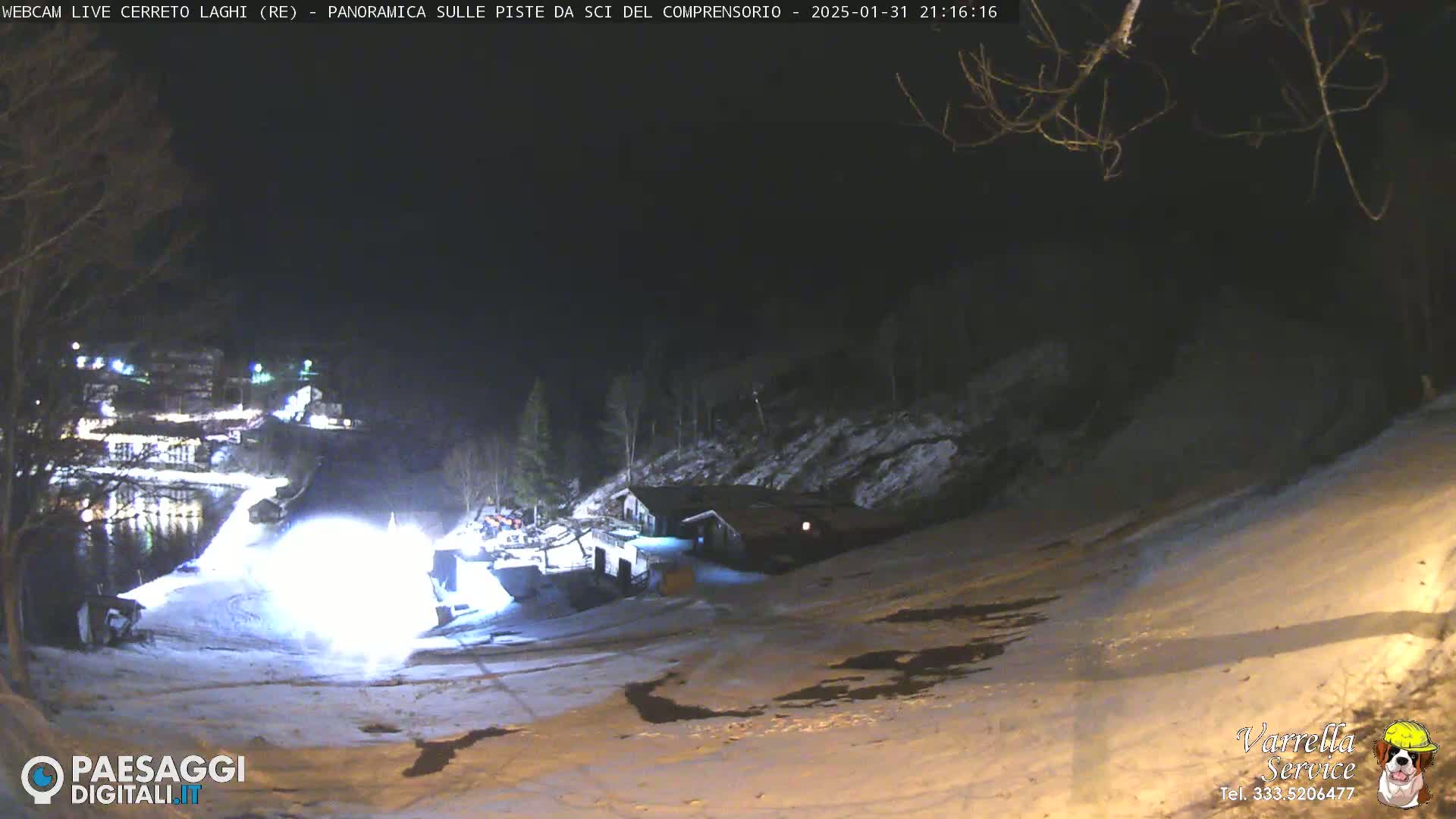 A snow-covered ski slope at night is illuminated by bright lights near a building, with a snow-grooming machine working on the lower part of the slope.