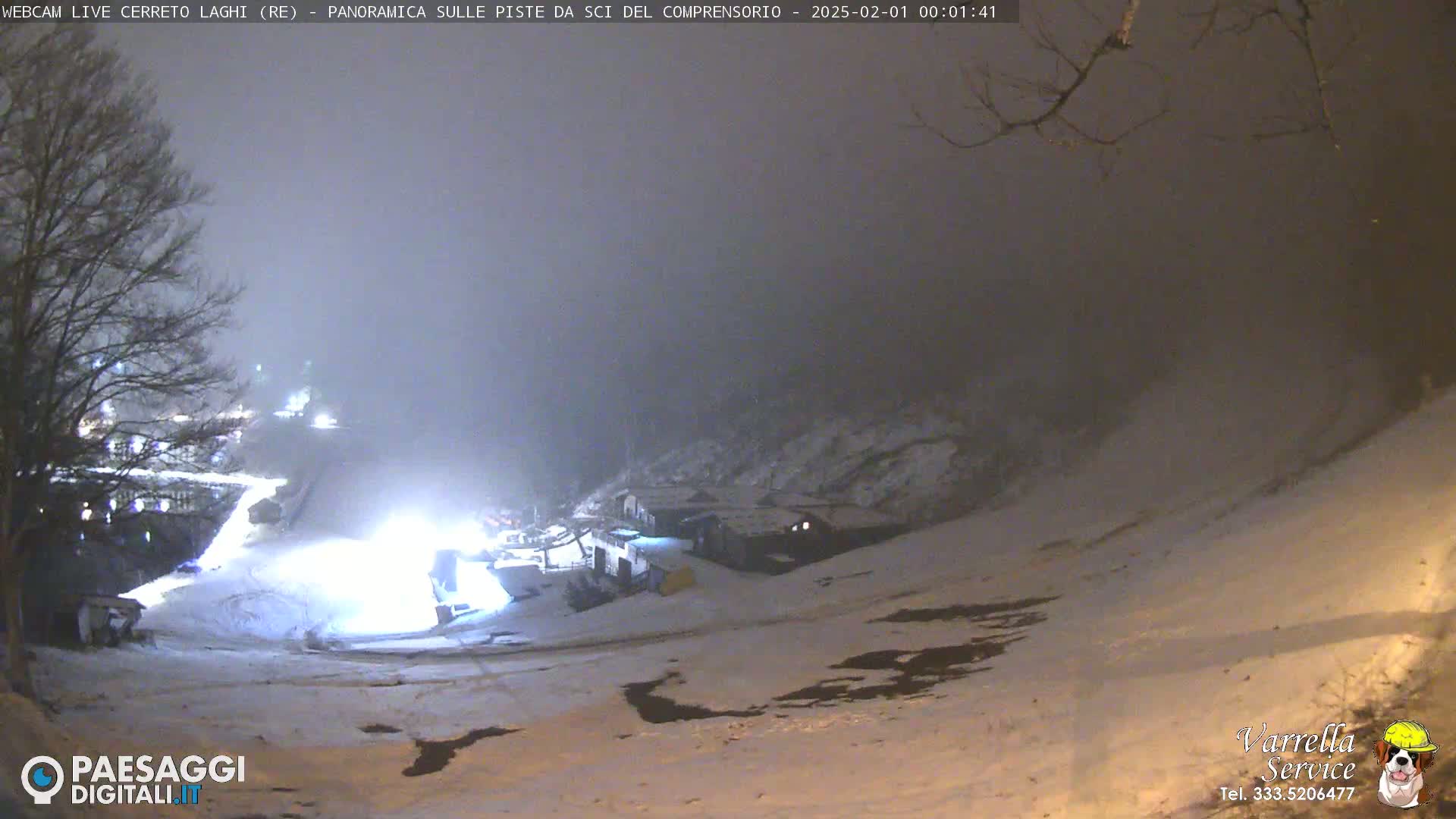 A snowy, nighttime scene shows a dimly lit ski slope with a few buildings and trees under a foggy sky.
