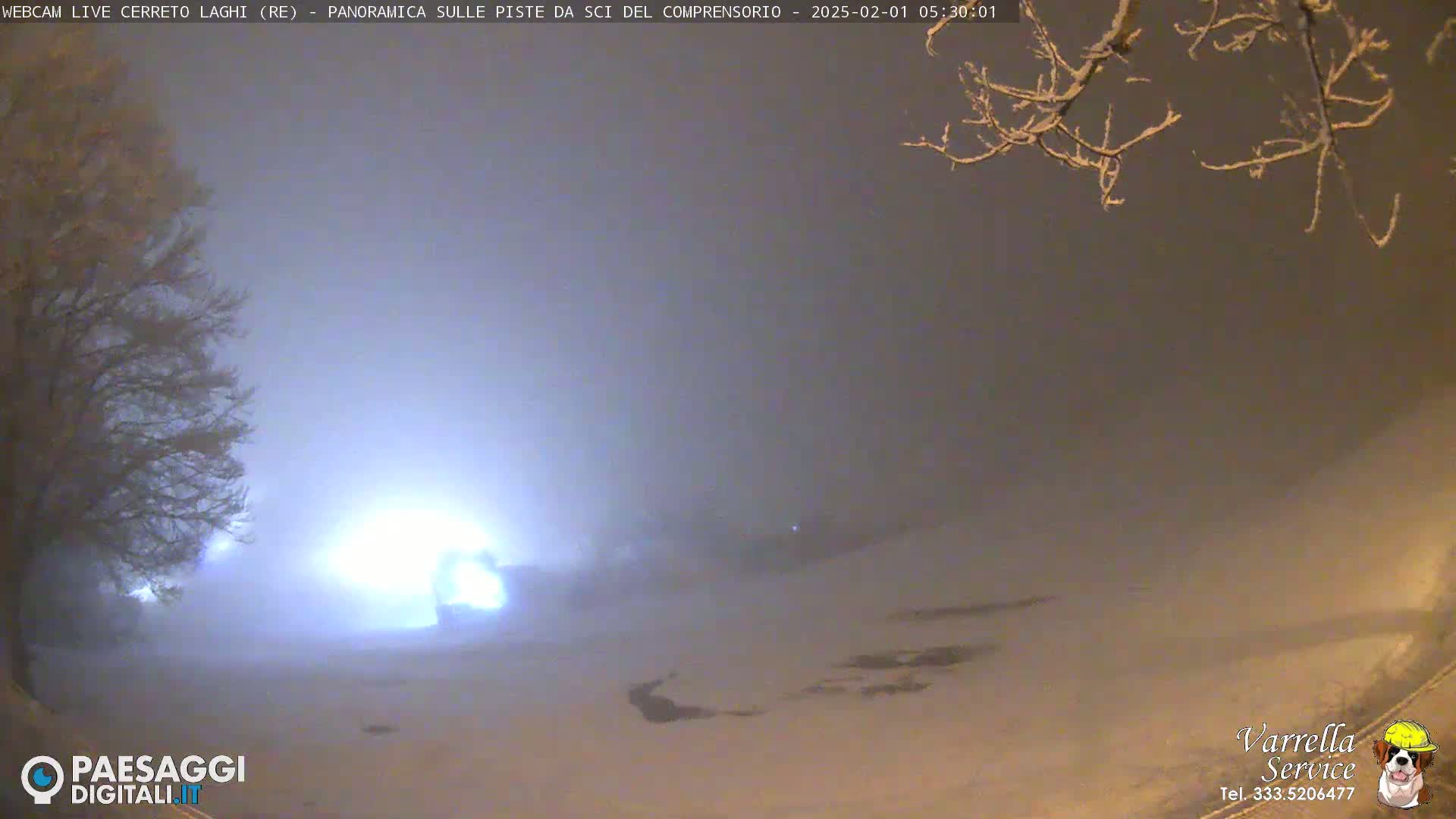 A snow-covered landscape is dimly lit at night under a hazy sky, with a few lights visible in the distance.