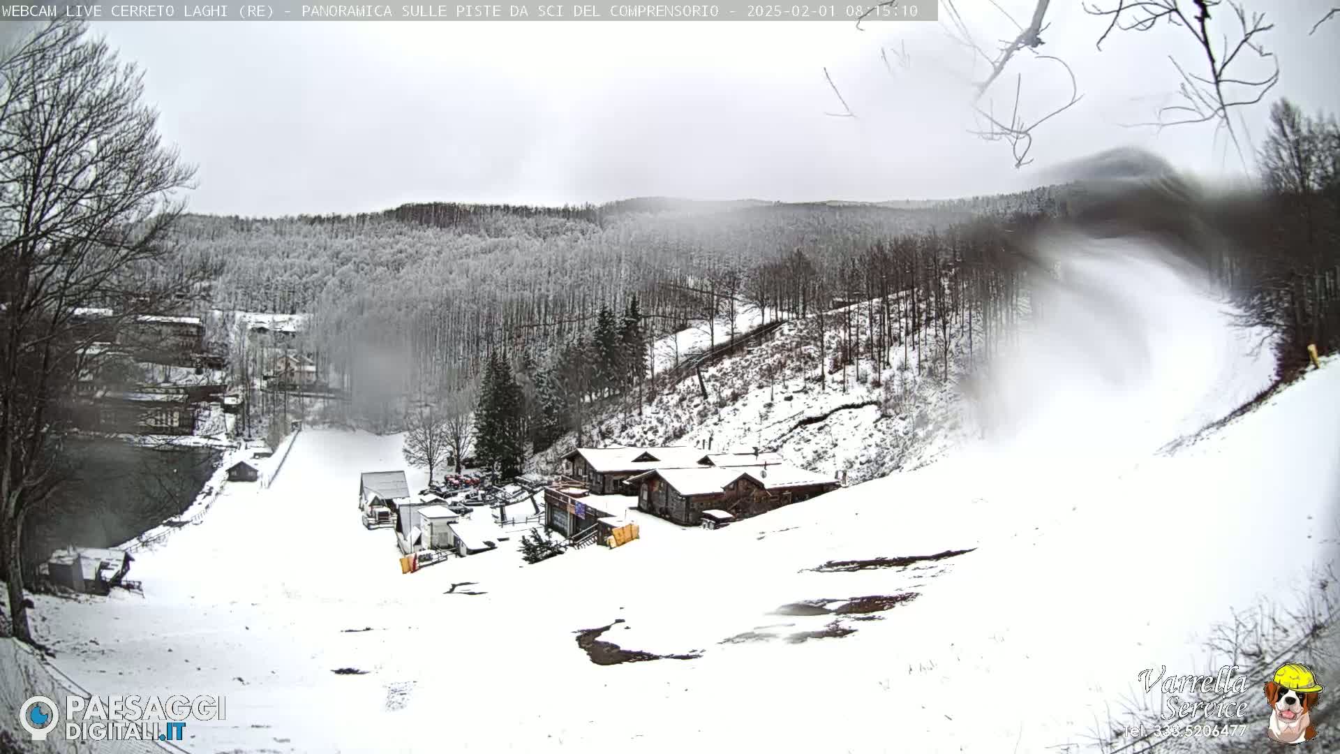 A snow-covered ski resort with several buildings, a snow-making machine operating, and a mostly cloudy sky.